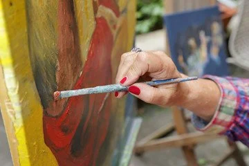 Close-up of a person painting a canvas with a paintbrush, wearing a plaid shirt and red nail polish.
