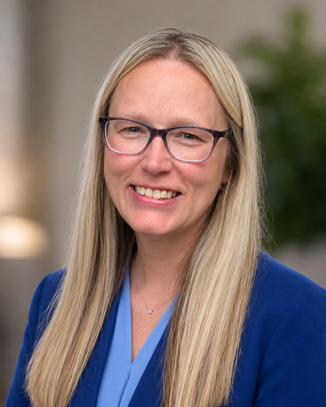 A woman with long blonde hair, glasses, and a blue blazer smiling at the camera.