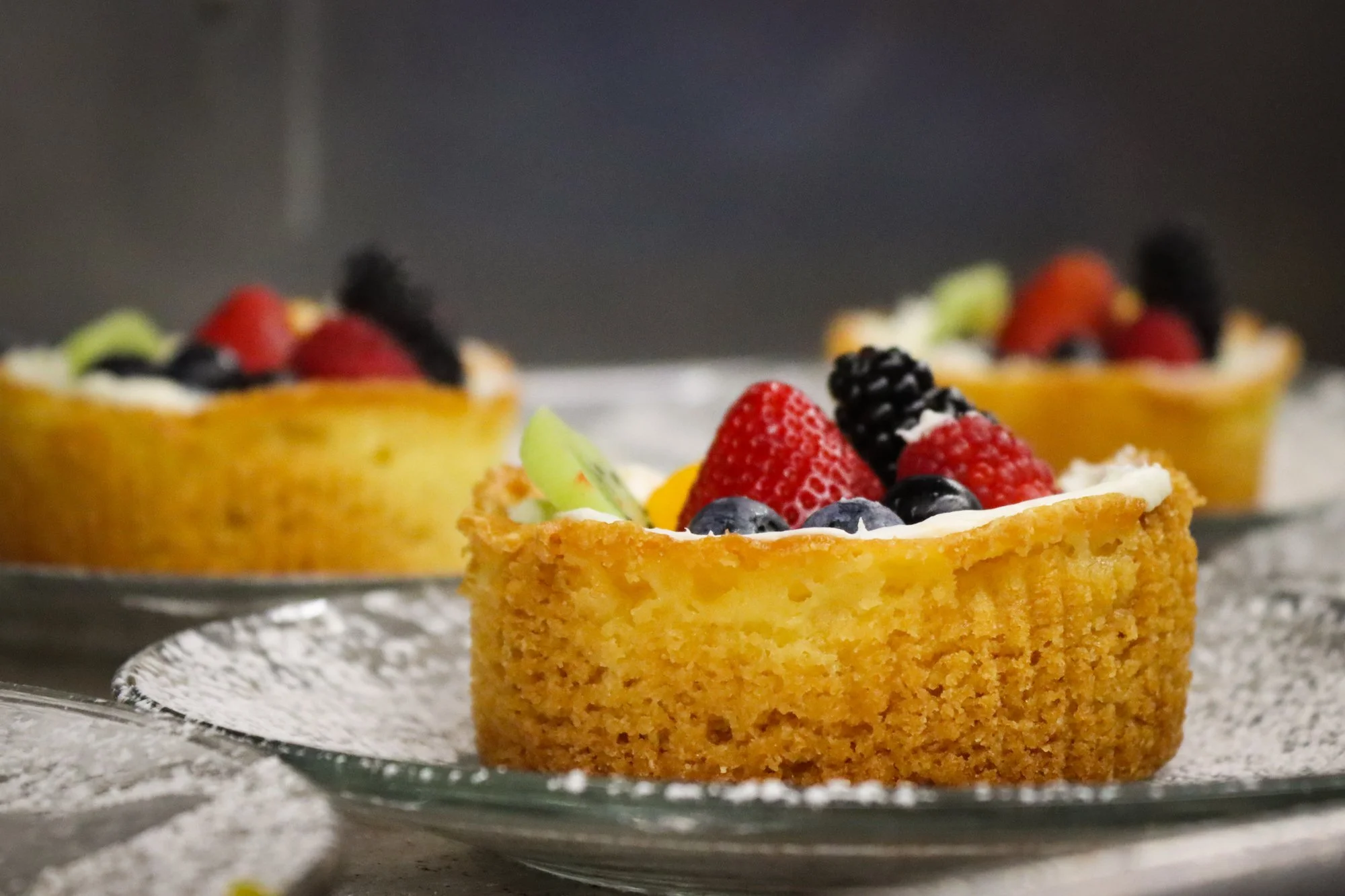 Close-up of a fruit tart with a golden crust, topped with strawberries, blackberries, blueberries, and green fruit slices. The tart is on a decorative glass dish with powdered sugar around it, on a gray surface.