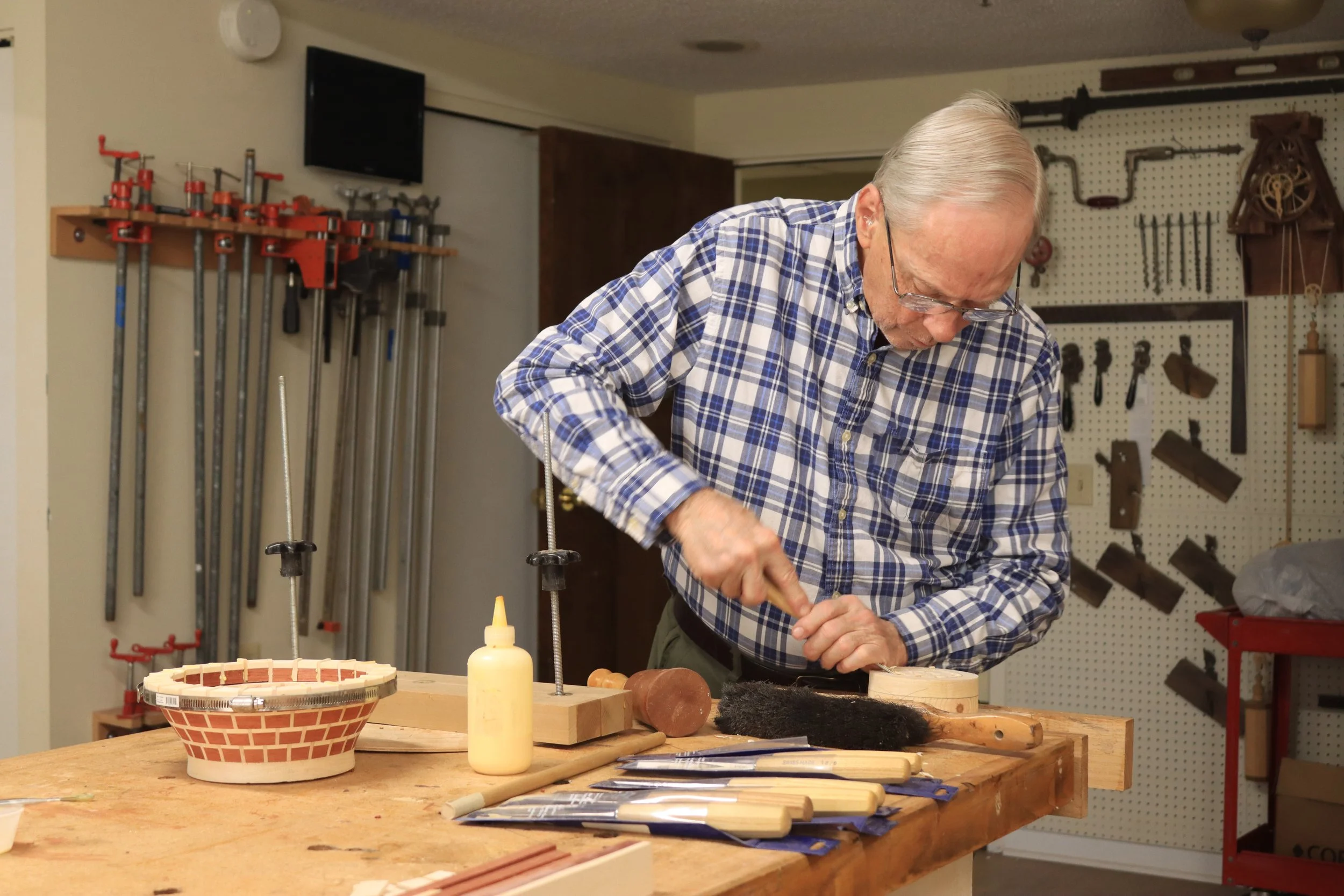 An elderly man with gray hair and glasses working at a woodworking bench in a workshop. The wall behind him has various tools mounted on a pegboard. The workbench has woodworking tools, a yellow squeeze bottle, and a basket.