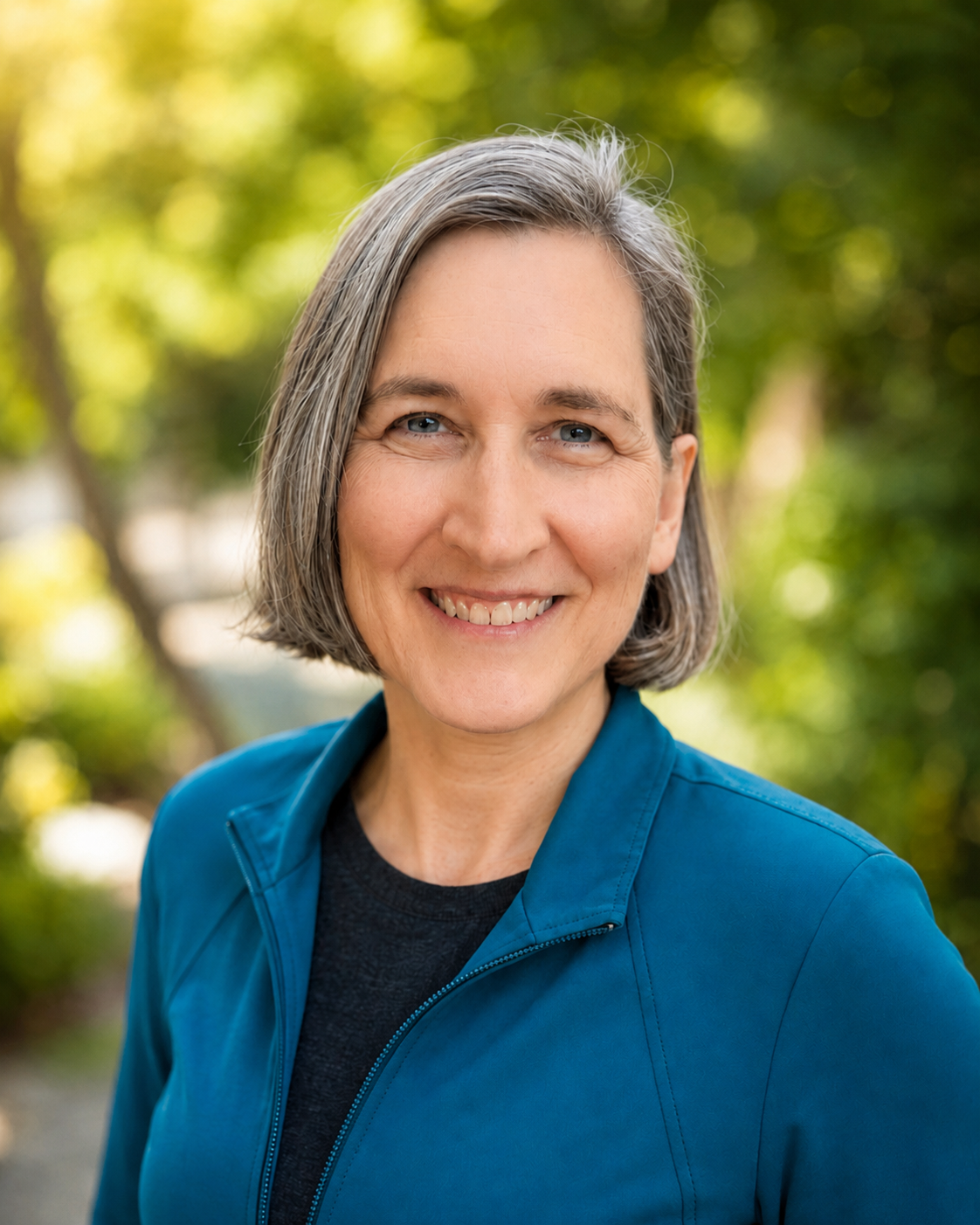 A middle-aged woman with short gray hair, wearing a blue jacket and black shirt, smiling outdoors with a blurred green foliage background.