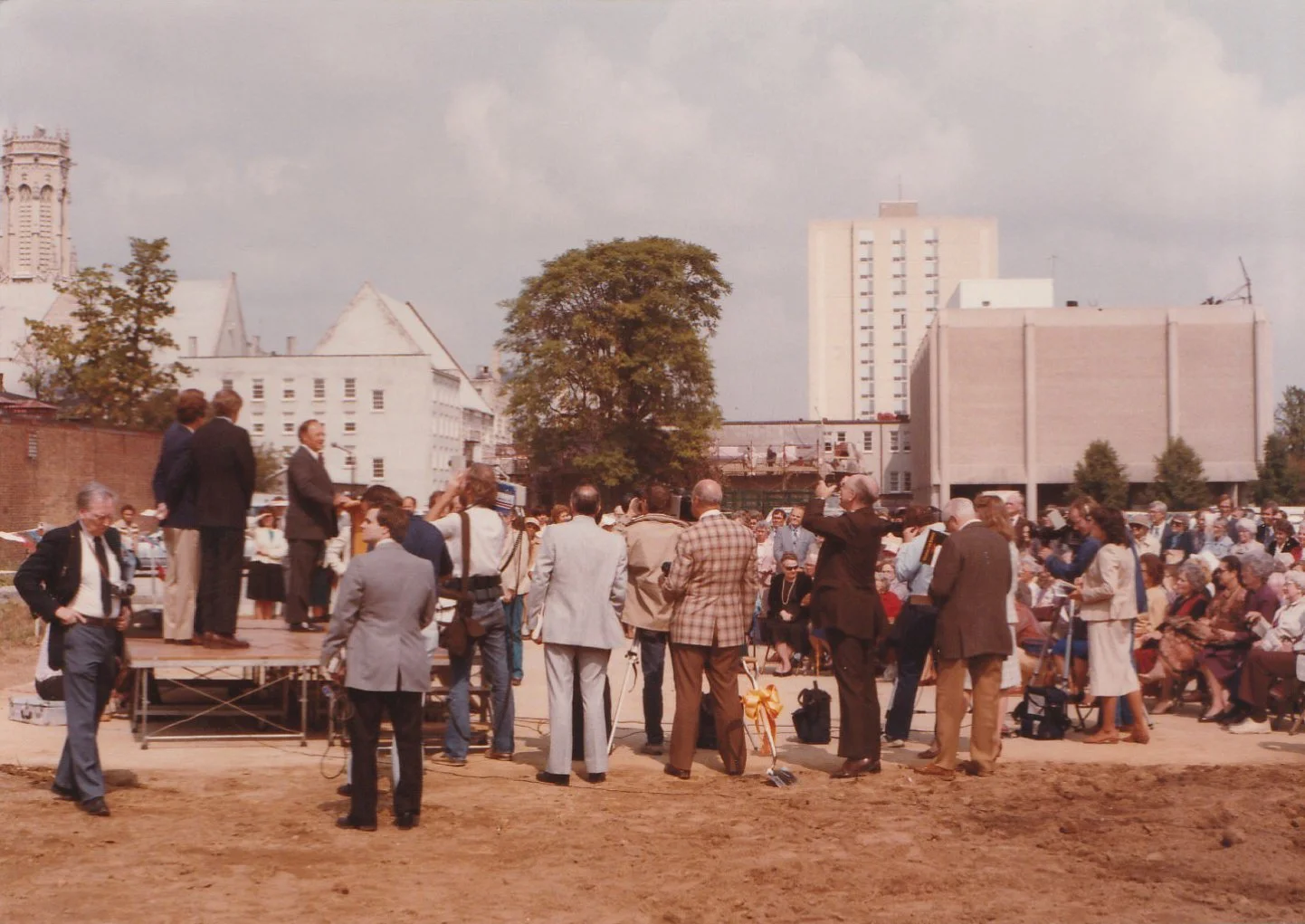 A group of people gathered outdoors for a formal event, with some standing on a small stage, others seated, in an urban setting with buildings and trees in the background.