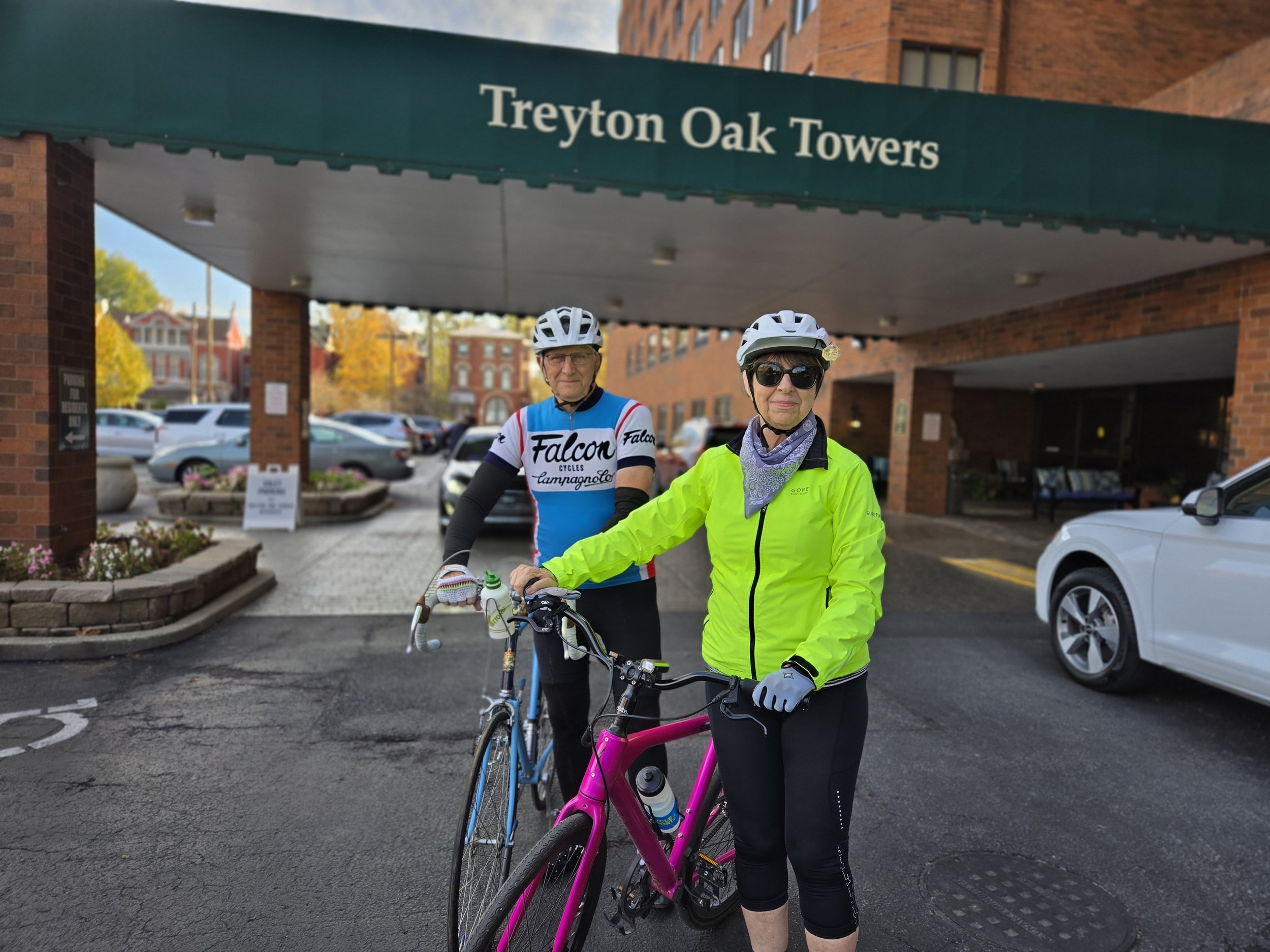 Two women wearing helmets and cycling gear posing with bicycles in a parking lot outside a building with a sign that reads 'Treyton Oak Towers'.
