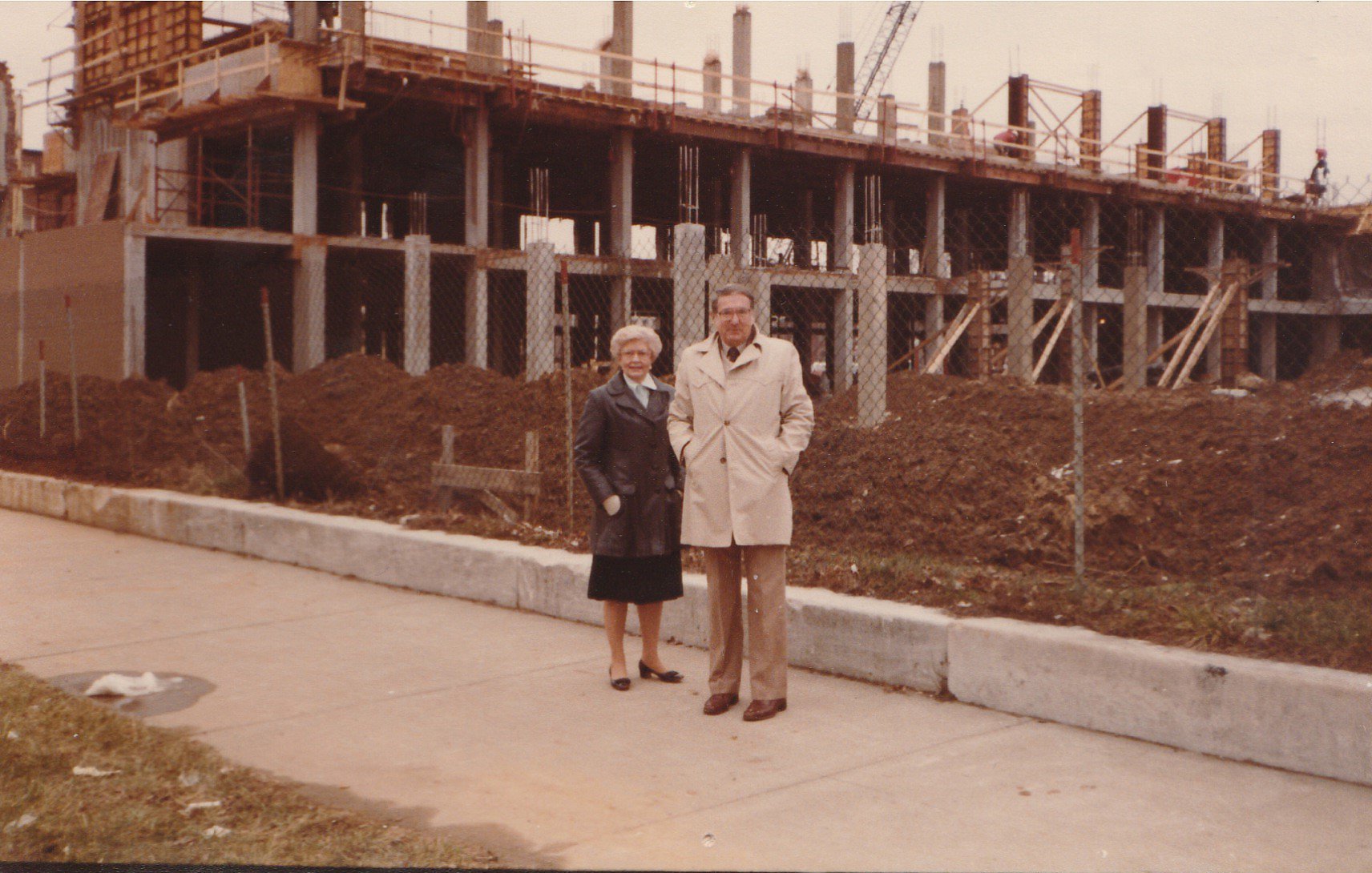 An older man and woman in vintage clothing standing on a sidewalk in front of a construction site with a partially built building and construction workers.