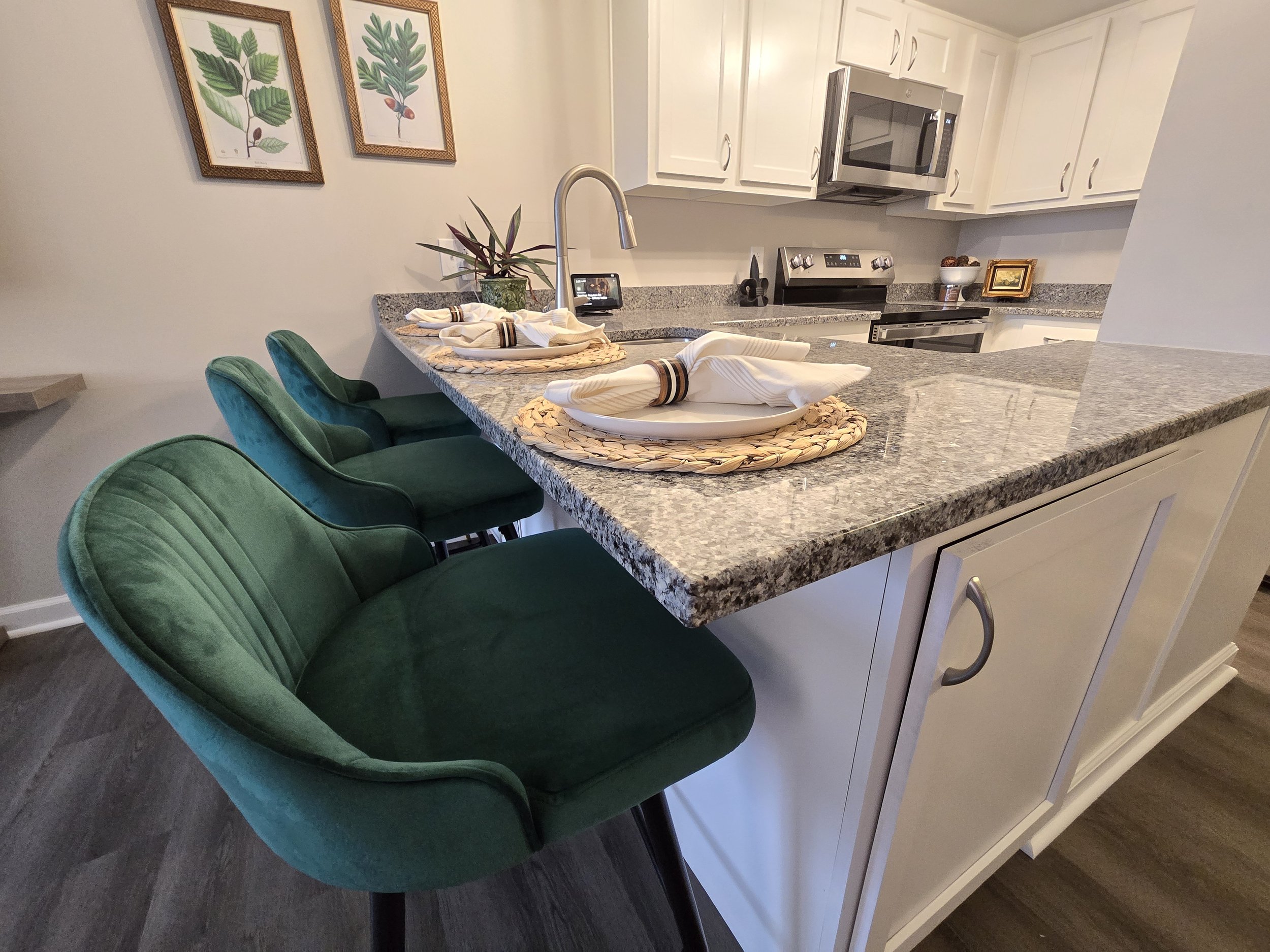 A kitchen with a granite countertop island, green upholstered chairs, and white cabinetry. Decorative art prints of plant illustrations hang on the wall, and there is a potted plant on the counter near a tablet. The kitchen also features stainless steel appliances, including a microwave and stove.