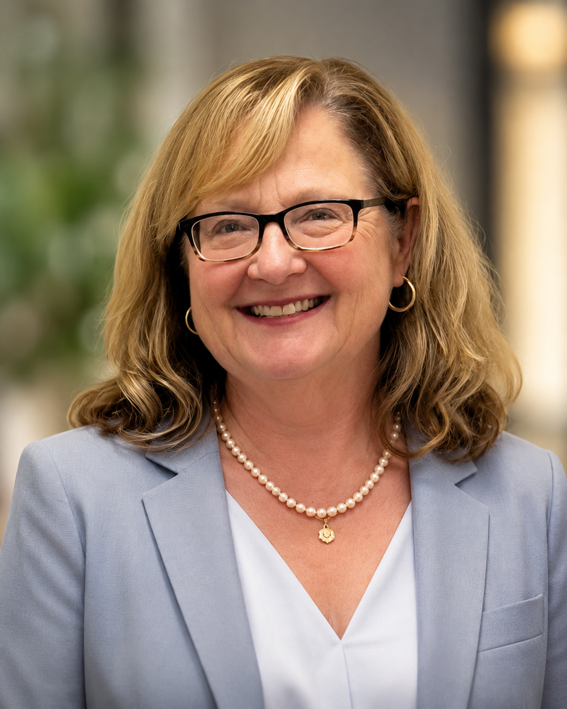 A smiling middle-aged woman with glasses and wavy shoulder-length hair, wearing a light blue blazer, a pearl necklace, and gold hoop earrings, standing indoors with blurred background.