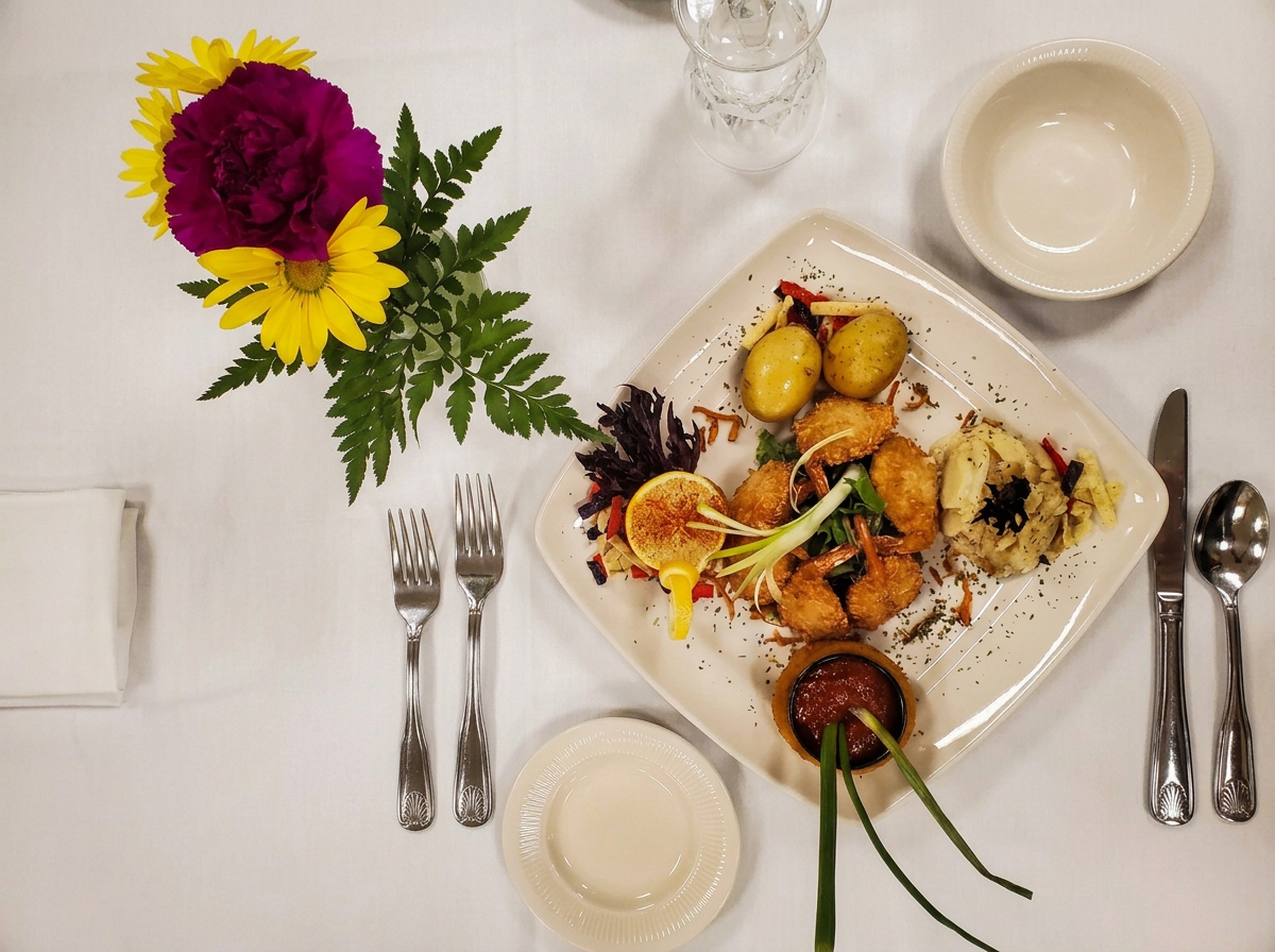 A table setting with a vase of colorful flowers, a plate of food, and utensils. The flowers include yellow daisies and a purple carnation. The food plate has potatoes, fried items, a lemon slice, and a small bowl of sauce. There are empty bowls and glassware on the table.
