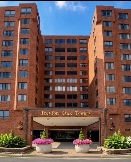 Front view of a tall red brick residential building with a sign reading "Treyton Oak Towers" at the entrance, featuring landscaped plants and flowers.