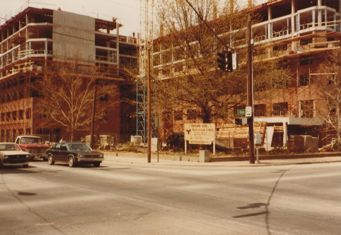 Construction site of a multi-story building with scaffolding, trees, and parked cars in the foreground, a sign indicating 'Future Site of FreyJa Oak Tower,' and traffic lights at an intersection.