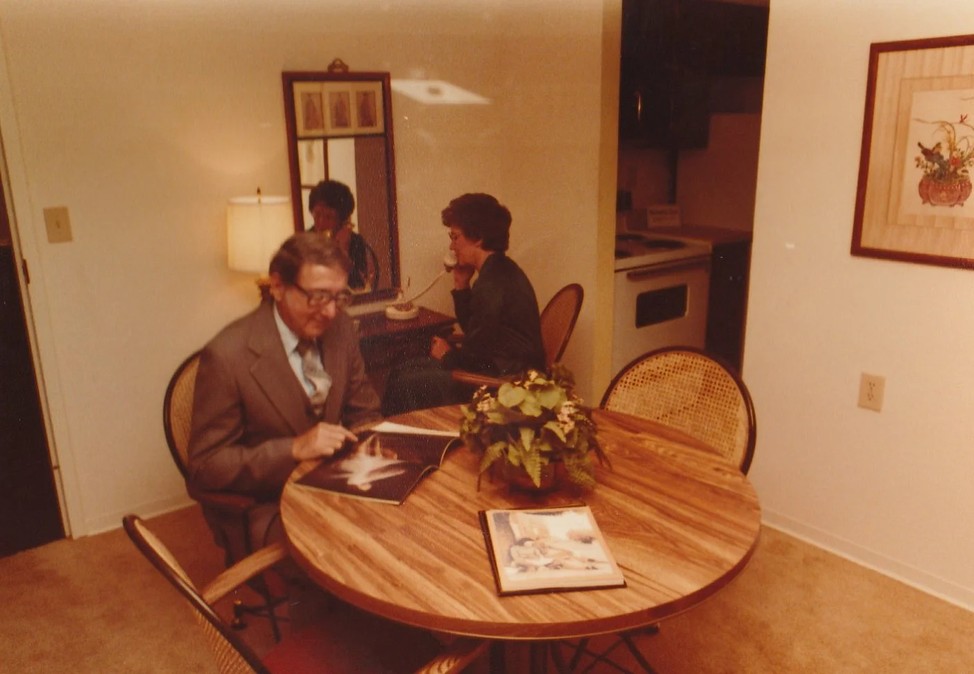 Vintage photo of three people in a dining area, one man in a suit sitting at a round wooden table looking at a magazine, two women are in the background, one talking on the phone and the other standing near a mirror, with a kitchen visible in the background.