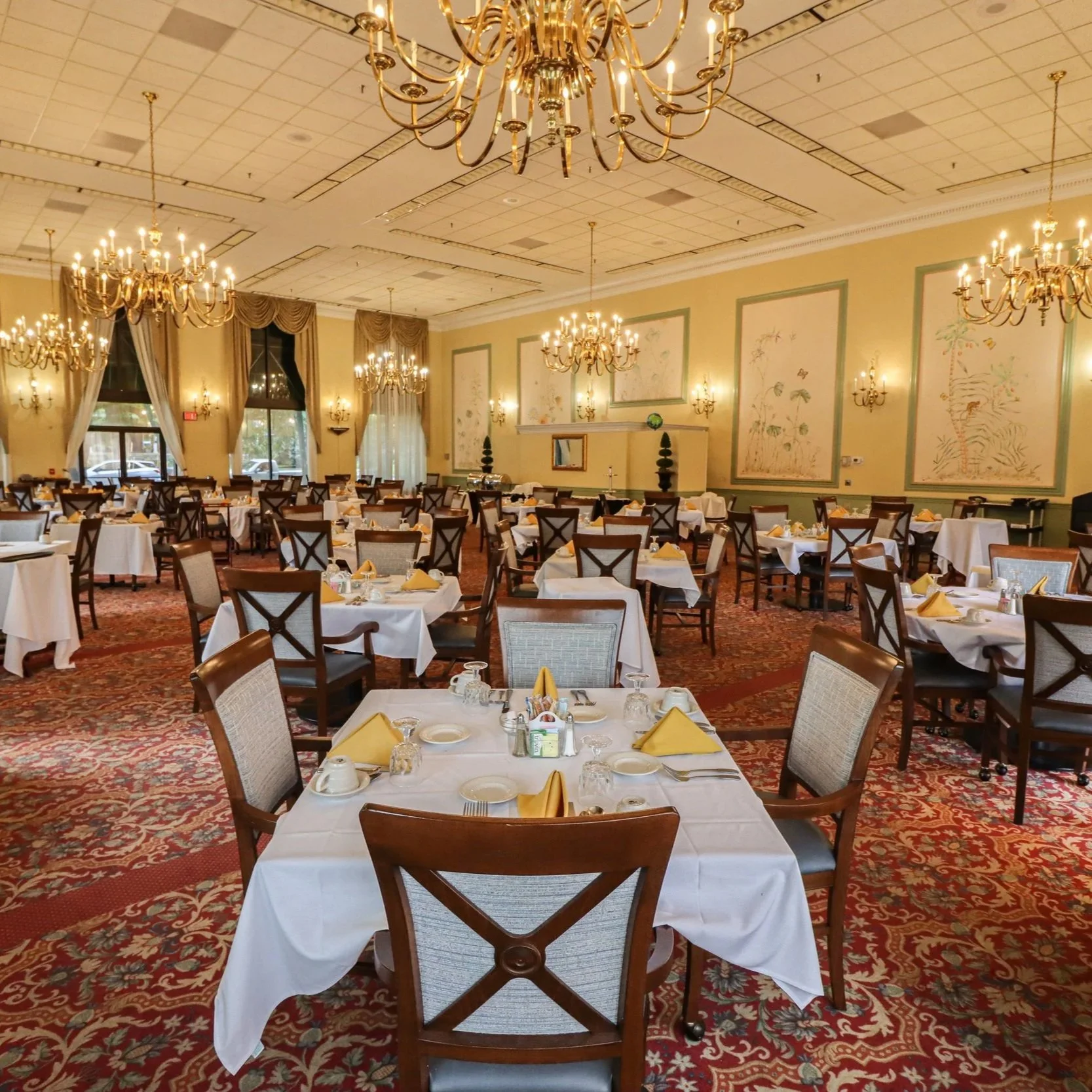 Elegant banquet hall with multiple round tables dressed with white tablecloths, yellow napkins, and place settings. The hall features large chandeliers, wall sconces, and framed artwork, with tall windows draped with curtains and patterned carpet flooring.