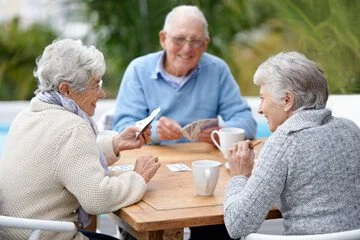 Three elderly women sitting at a wooden table outdoors, enjoying conversation. Two are looking at photographs, one is holding a cup of coffee or tea.