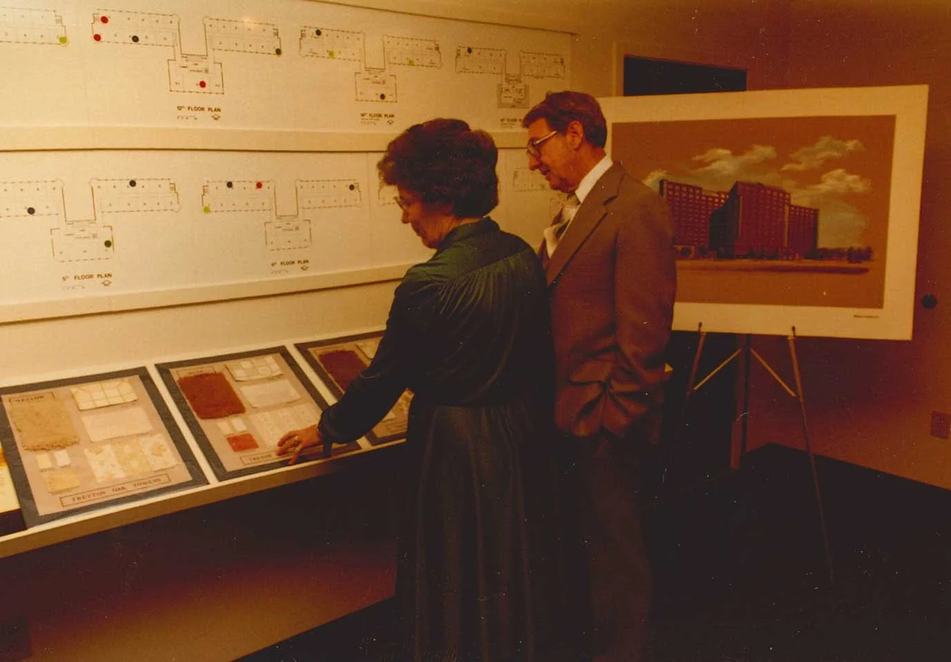 Two people, a woman and a man, are standing in front of display boards with architectural plans, drawings, and informational materials, likely at an exhibition or presentation. The woman is pointing at the materials, and there is a large painting of a building on an easel nearby.
