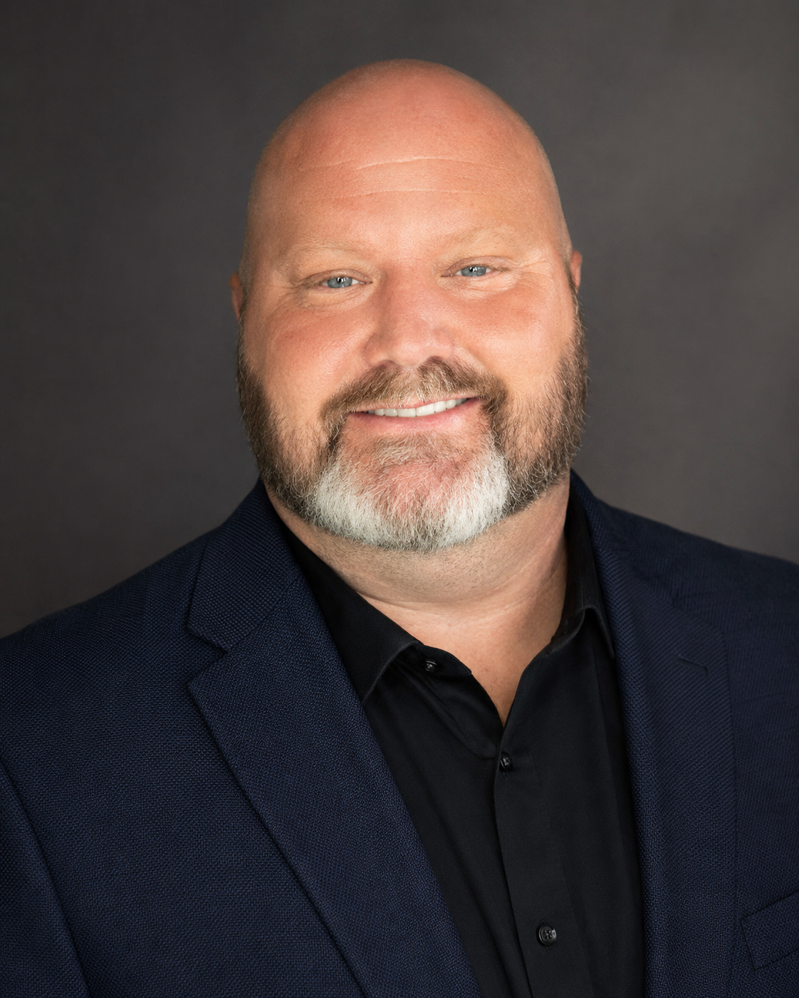 A headshot of a smiling middle-aged man with a beard and bald head, wearing a dark suit and black shirt, against a dark background.