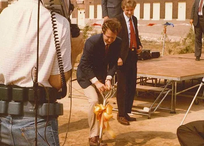 A man dressed in a suit with a large yellow bow on his bicycle, surrounded by people in formal attire at an outdoor event.