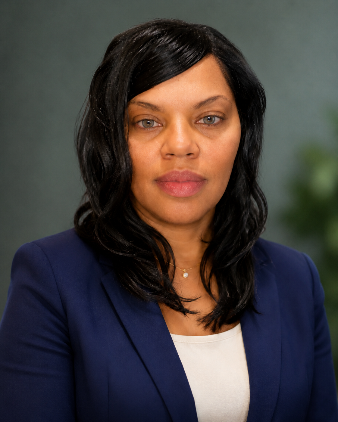 A woman with black wavy hair wearing a navy blazer over a white top, looking directly at the camera with a neutral background.