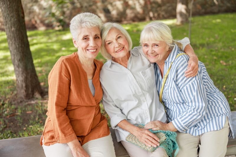 Three elderly women sitting close together on a park bench, smiling and embracing each other outdoors.