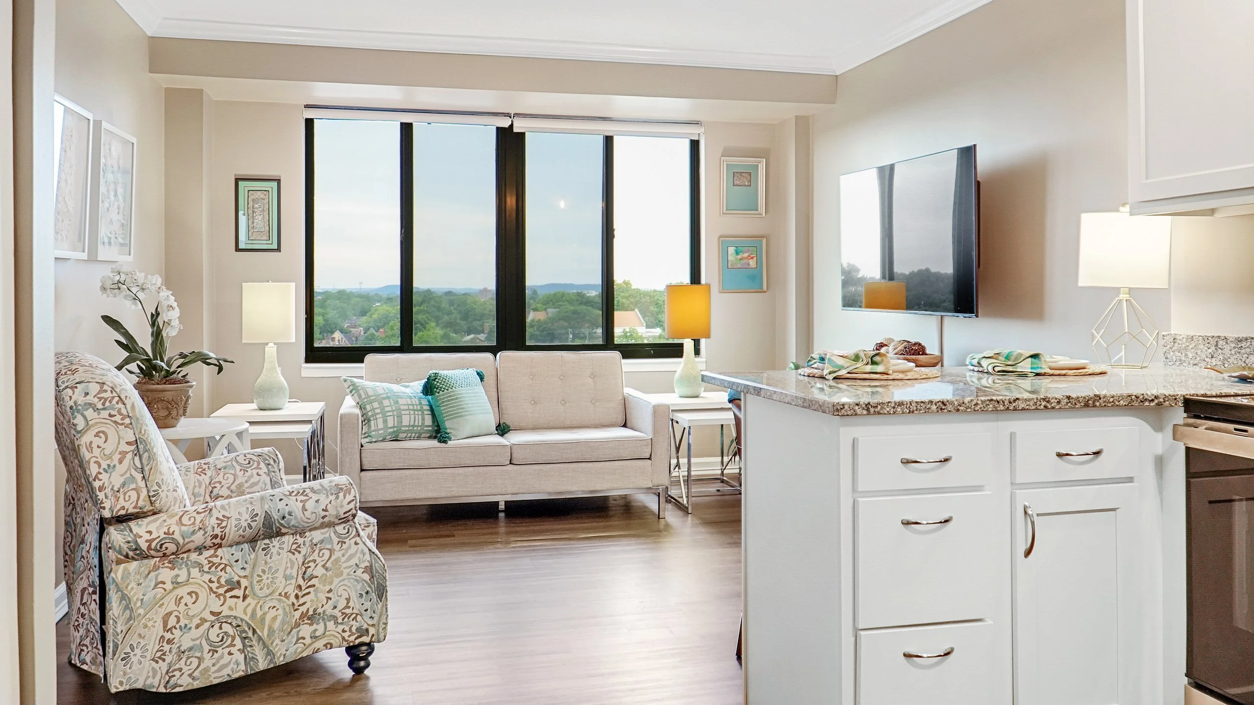 Living room with large windows, beige and patterned armchair, white sofa with green and white pillows, side tables with lamps, television on wall, and kitchen counter in the foreground.