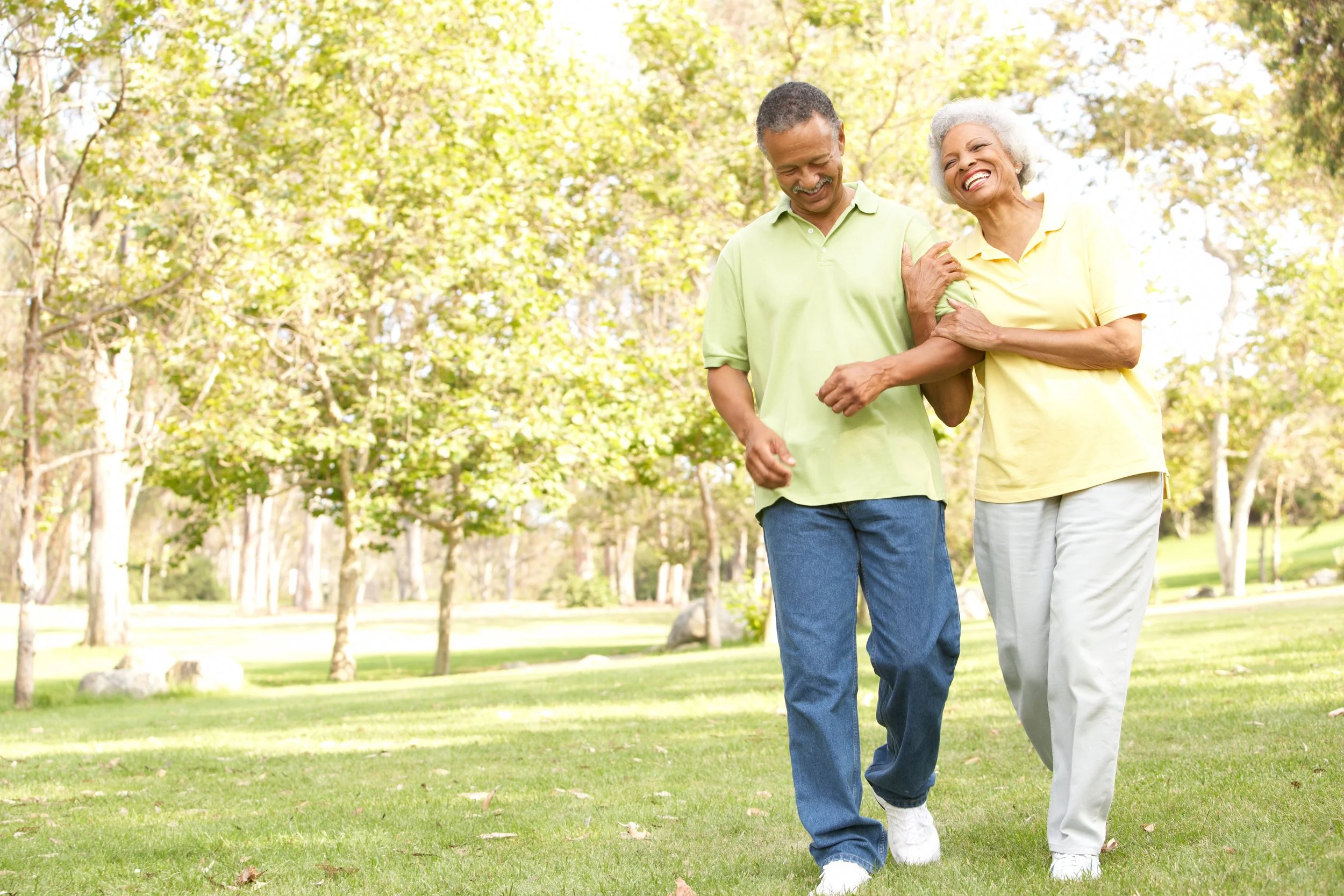 An elderly woman and a middle-aged man smiling and walking together in a park surrounded by green trees.