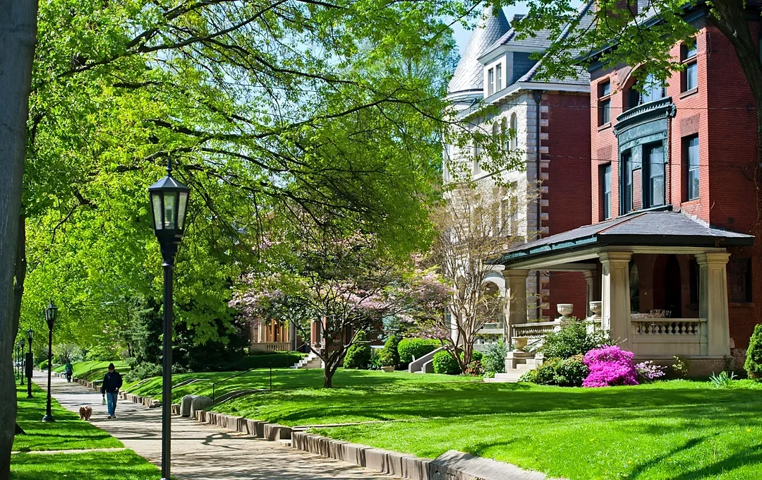 A peaceful residential neighborhood with lush green grass, trees, a sidewalk, and historic brick houses with porches, under a clear blue sky.