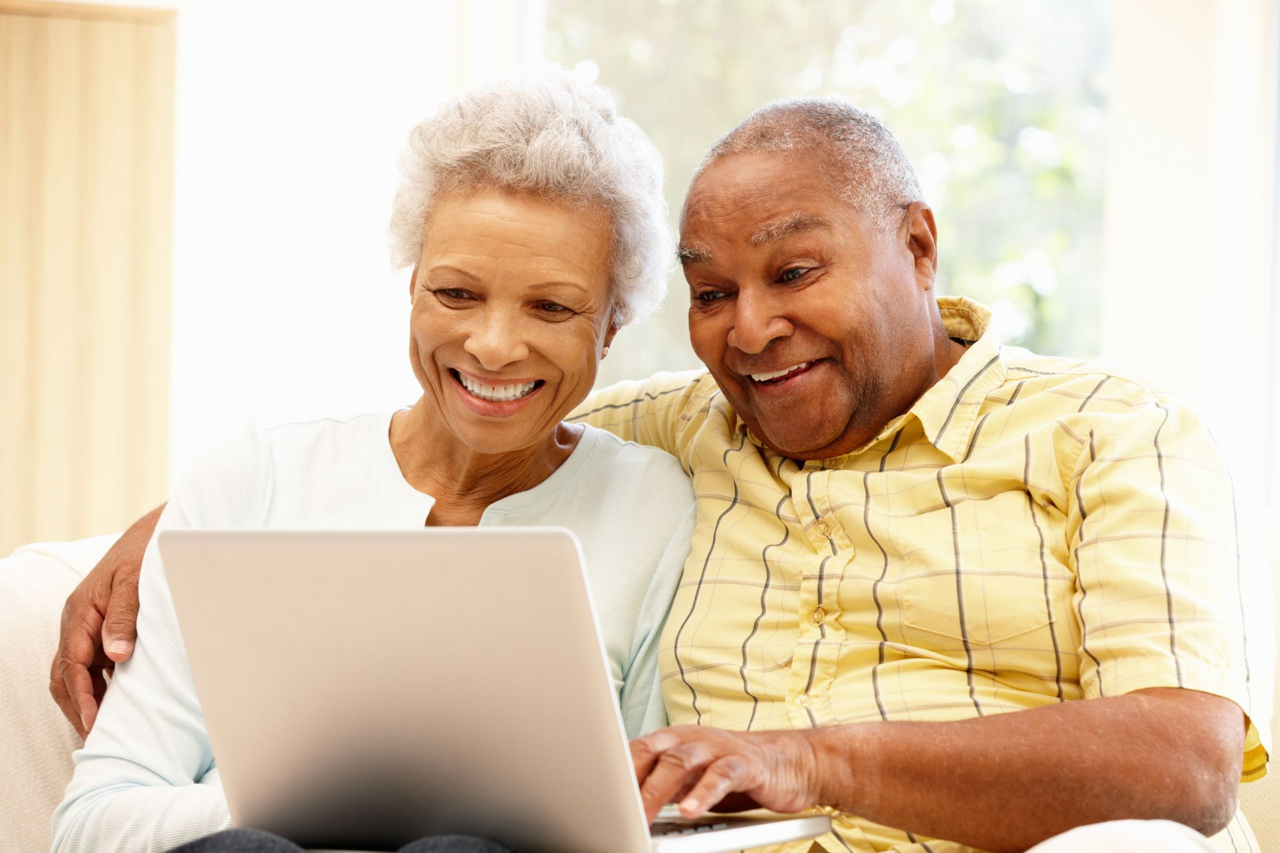 An elderly couple sitting close together on a beige sofa, looking at a laptop screen and smiling, with sunlight streaming through a window behind them.