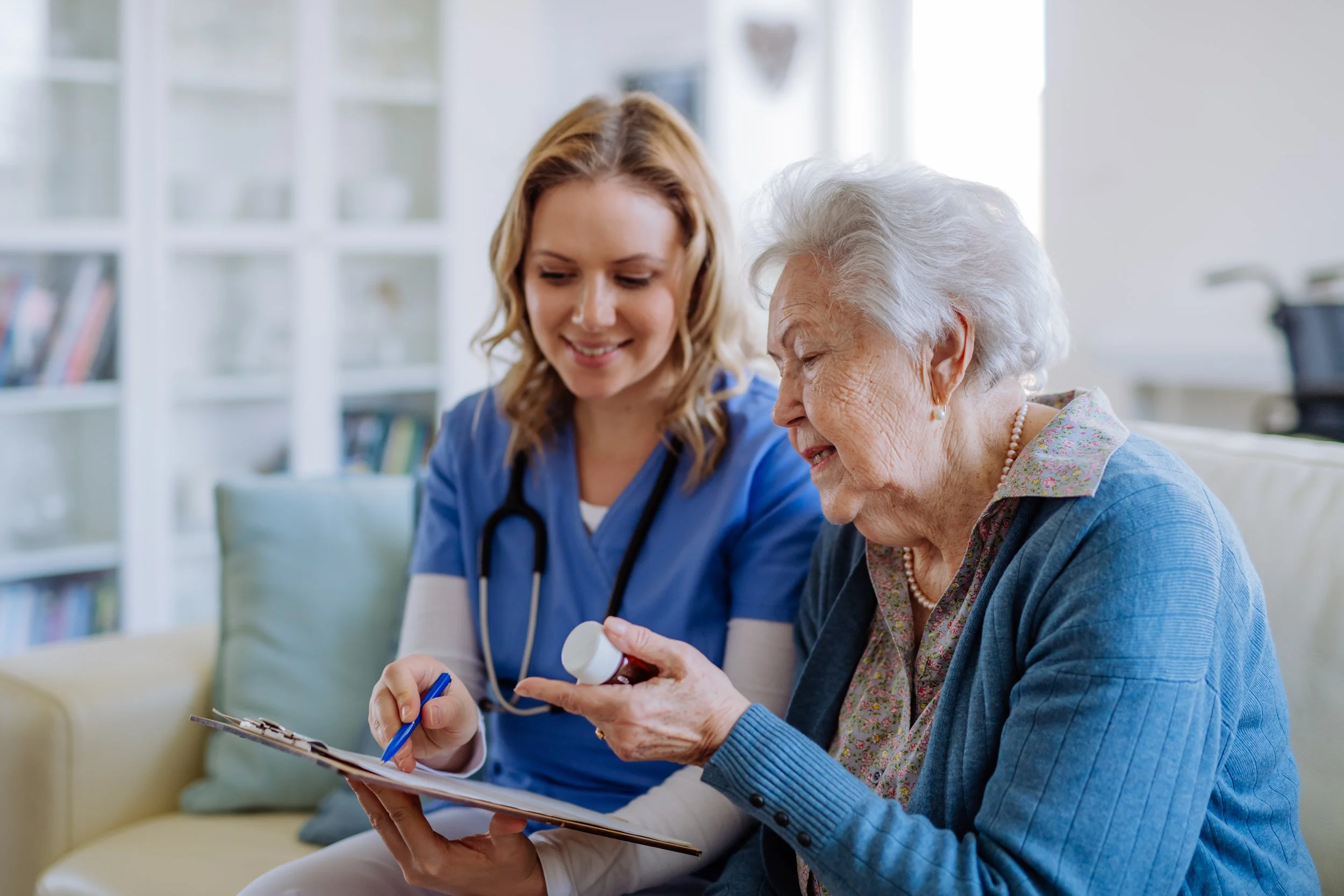 A young female healthcare professional with a stethoscope around her neck assisting an elderly woman with a medication bottle in a bright living room.