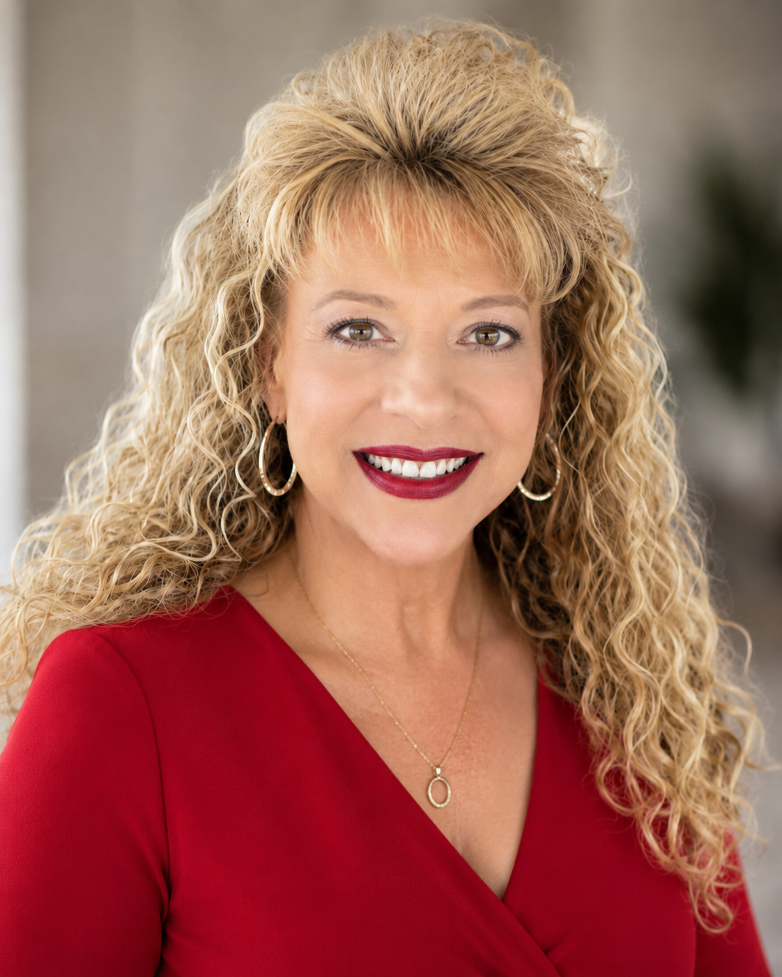 A woman with blonde curly hair wearing a red top, gold hoop earrings, a gold necklace, and smiling.
