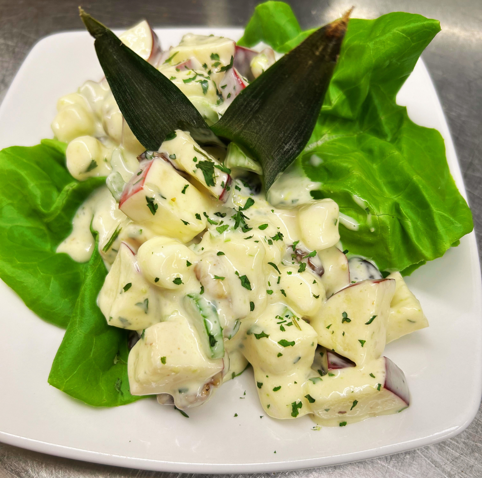 A plate of potato salad with chopped red onions, herbs, and a creamy dressing, garnished with parsley and served on a bed of leafy greens.