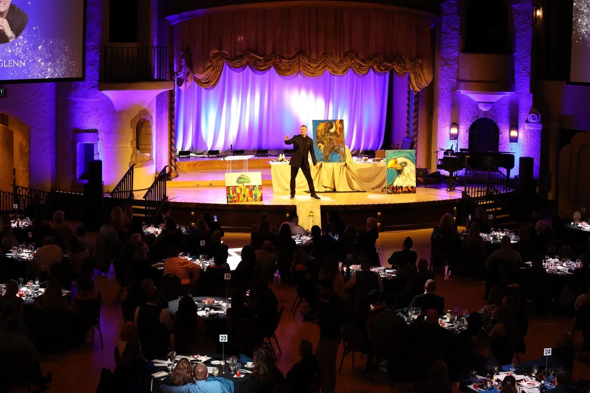 A stage with purple lighting and a closed curtain, featuring a man in a black suit giving a presentation or speech, surrounded by colorful paintings of animals including an eagle and a lion, with an audience seated at tables in an elegant venue.