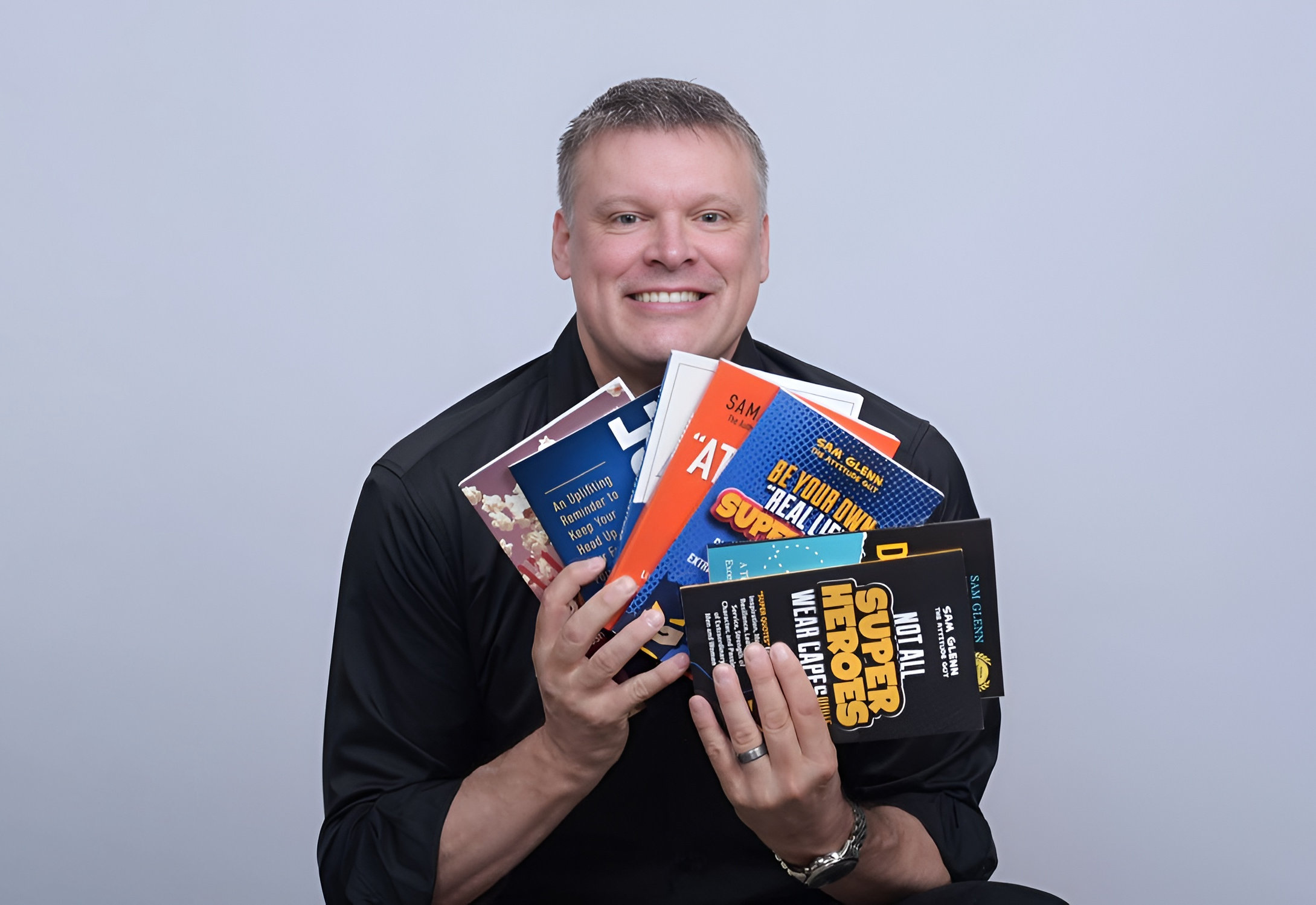 A man smiling and holding multiple colorful books or magazines in front of a plain light gray background.