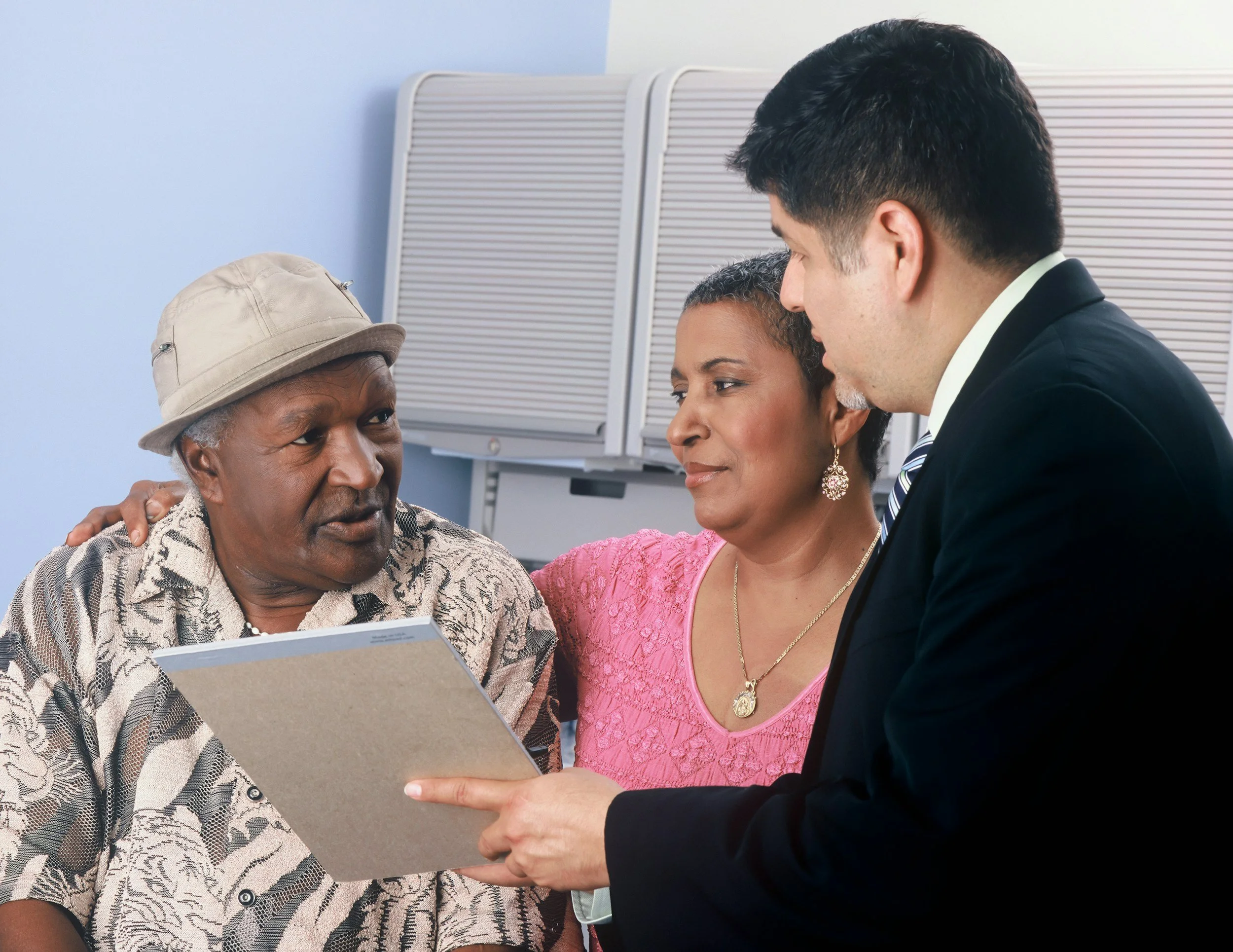 Three adults, two men and one woman, are engaged in a discussion, looking at a gray folder, in an indoor office setting.