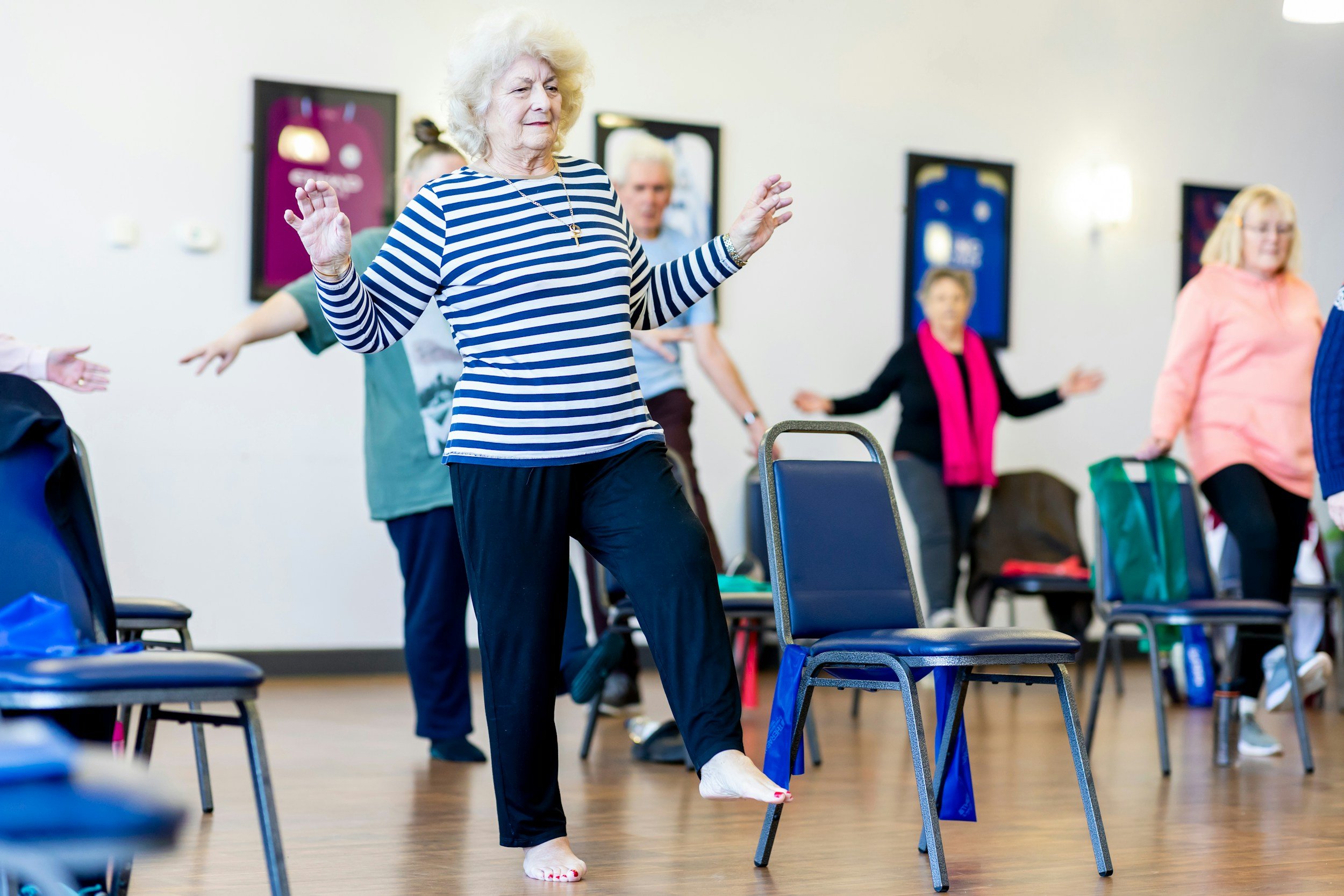 Older woman participating in a seated exercise class, balancing on one leg with arms raised, in a room with chairs and other participants exercising in the background.