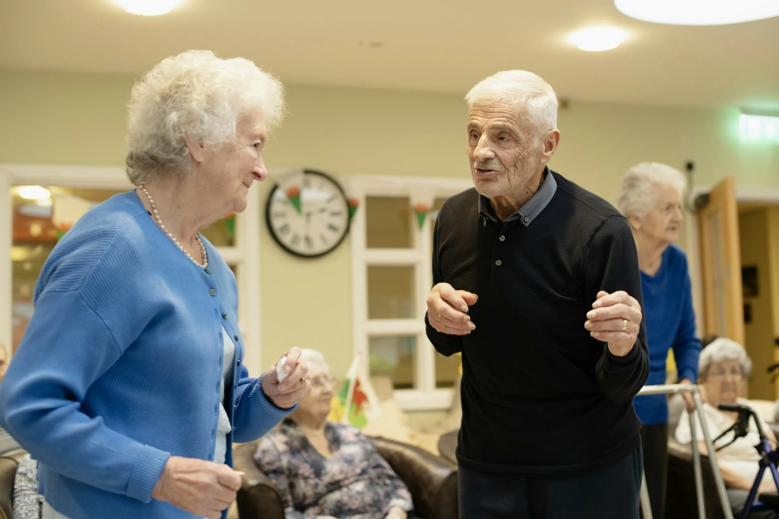 An elderly man and woman engaging in conversation at a social gathering in a senior living facility, with other elderly residents in the background.