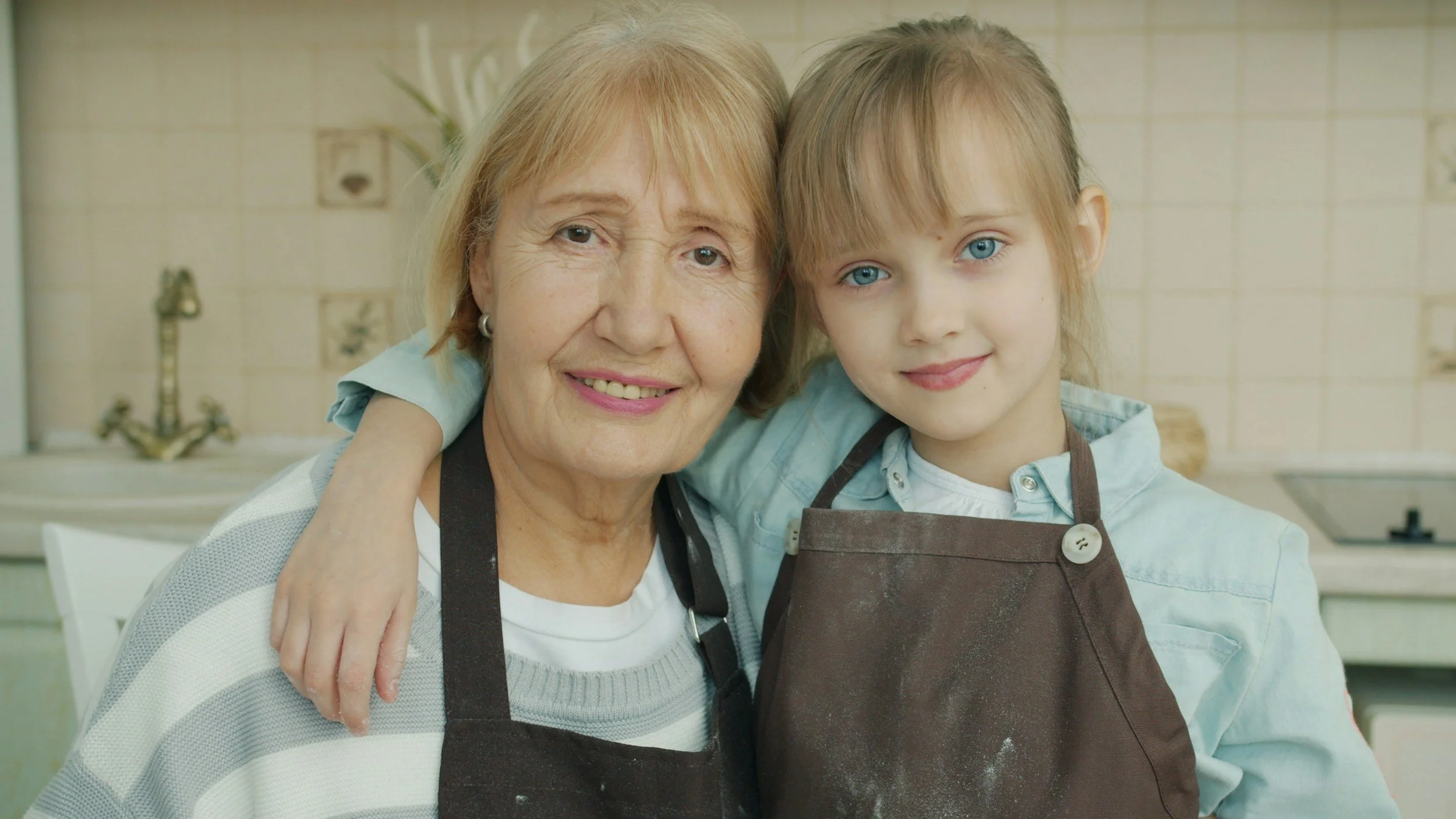 A grandmother and a young girl with blue eyes smiling and hugging each other in a kitchen, both wearing aprons.