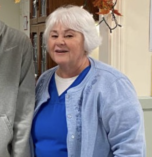 An elderly woman with short white hair wearing a blue shirt and a light blue cardigan, smiling.