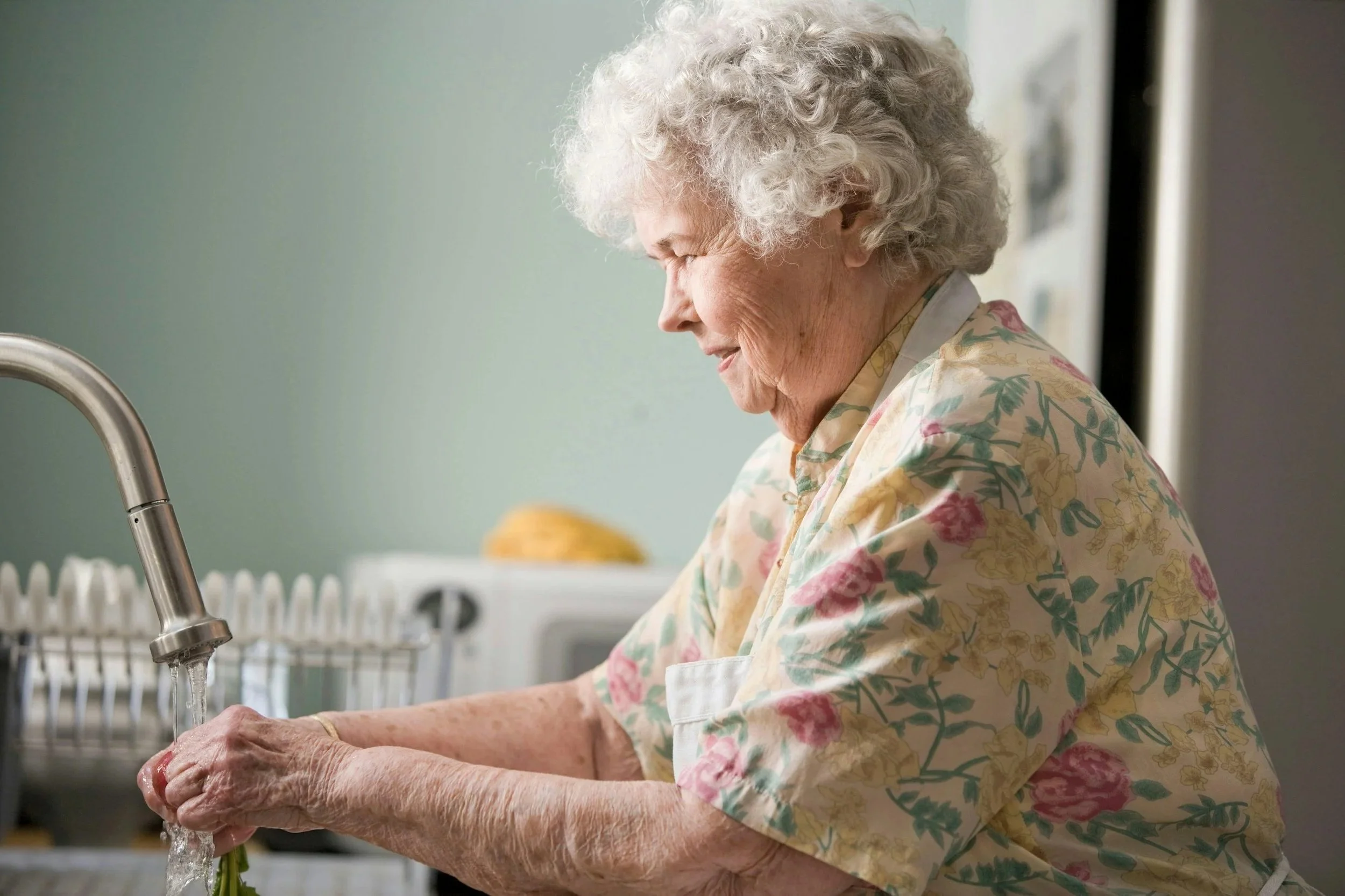 An elderly woman with curly gray hair washing vegetables in the kitchen under a running sink faucet.