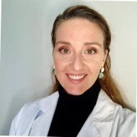 Portrait of a young woman with brown hair, wearing a white lab coat and a black top, smiling at the camera.