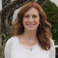 A woman with shoulder-length red hair, wearing a white top, standing outdoors with greenery in the background and smiling.