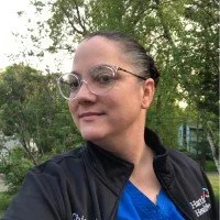 A woman with dark hair slicked back, wearing glasses and a dark jacket, standing outdoors in front of green trees.