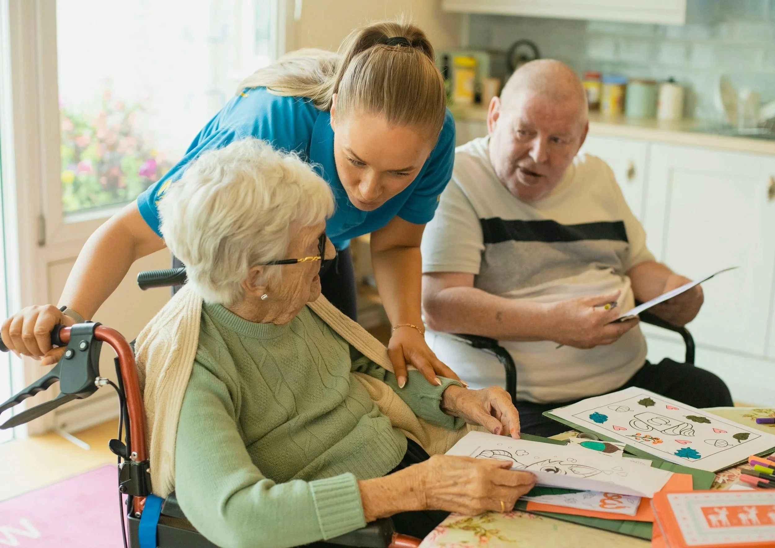 An elderly woman in a wheelchair looks at coloring pages on a table, accompanied by a caregiver leaning in to assist, and an elderly man sitting nearby with coloring pages. The scene takes place in a well-lit room with a kitchen in the background.