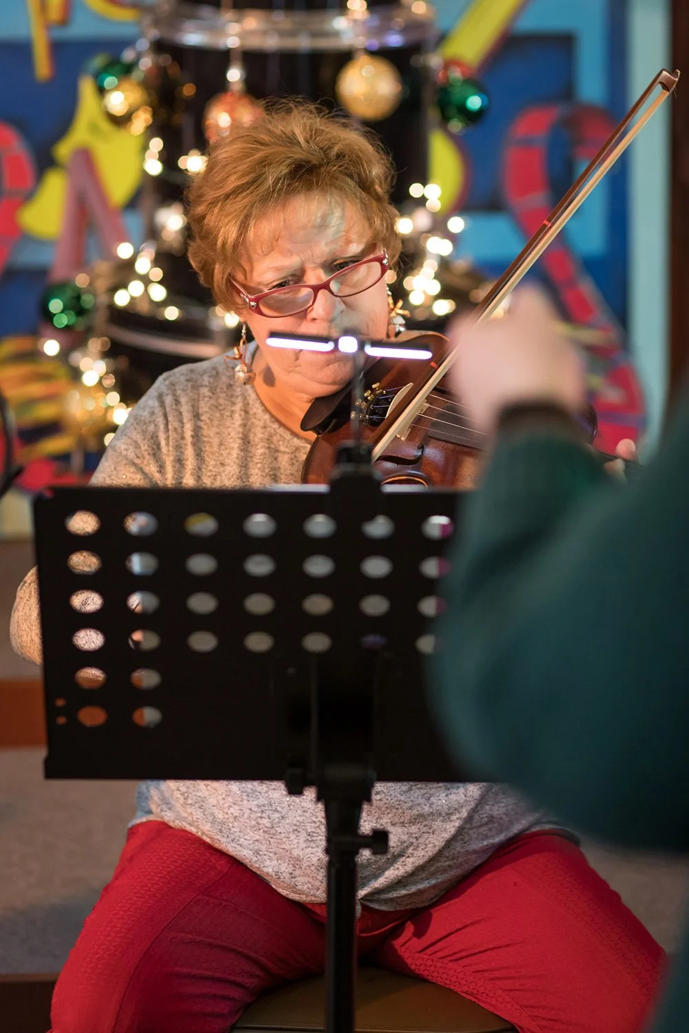A woman with short hair, glasses, and earrings playing the violin, focusing intently on sheet music, with Christmas decorations including ornaments and lights in the background.