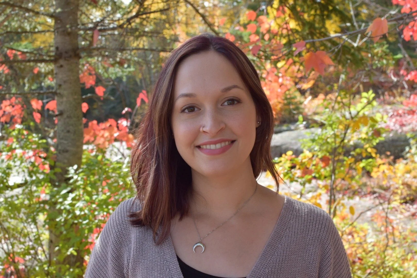 A woman smiling outdoors during fall, with colorful autumn leaves and trees in the background.
