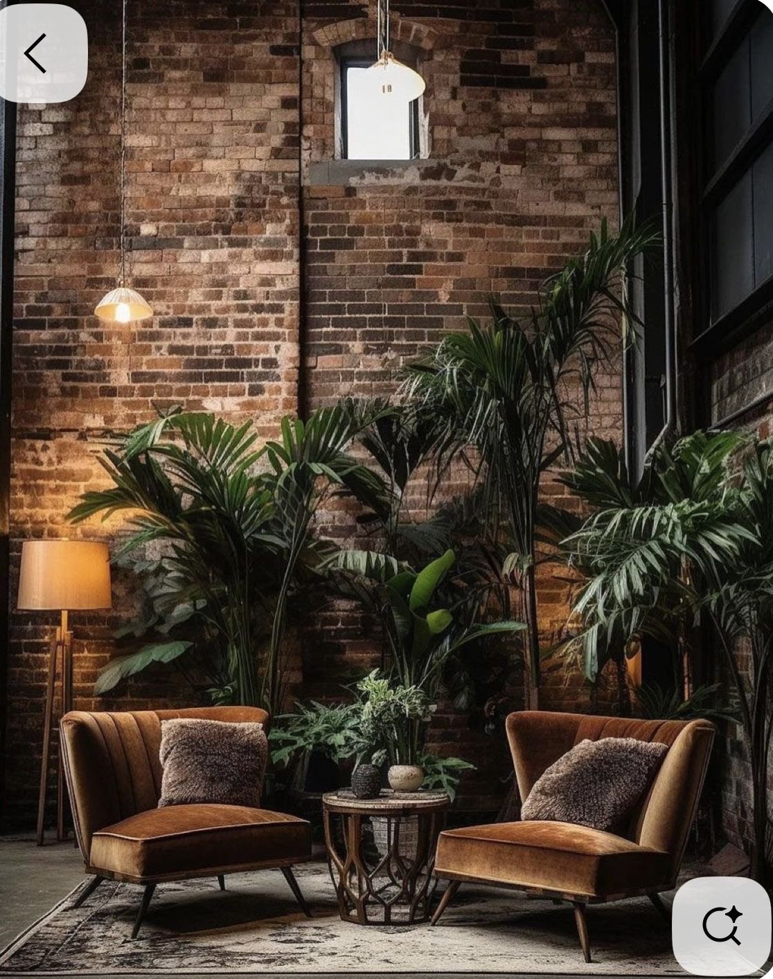Two vintage-style orange velvet armchairs with fur pillows, a small round wooden table with potted plants, large green leafy plants in the background, a brick wall, a tall floor lamp, a standing lamp, hanging pendant lights, and a rug.