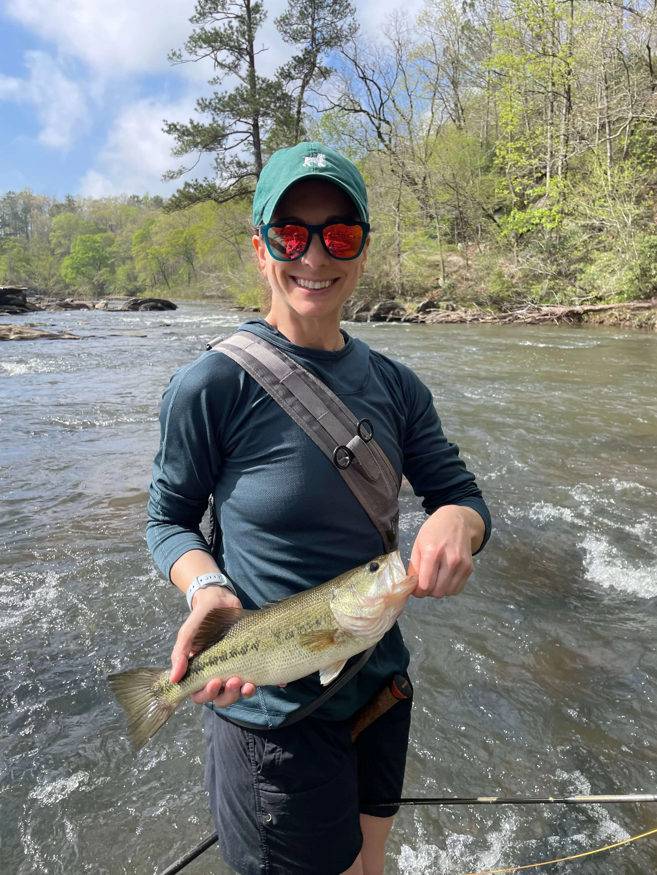 Person wearing sunglasses, hat, and outdoor clothing holding a largemouth bass fish in a river with trees and blue sky in the background.