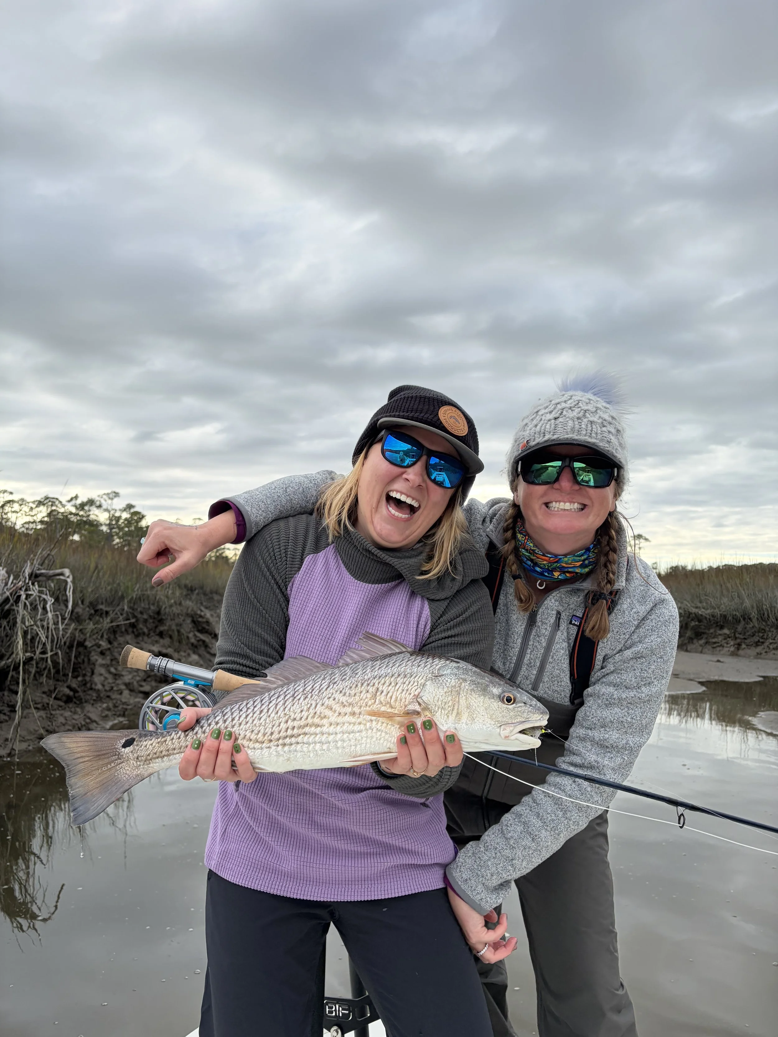 Two women wearing sunglasses and warm clothing are smiling and holding a large fish they caught while on a boat or near water, with a cloudy sky and marshy land in the background.