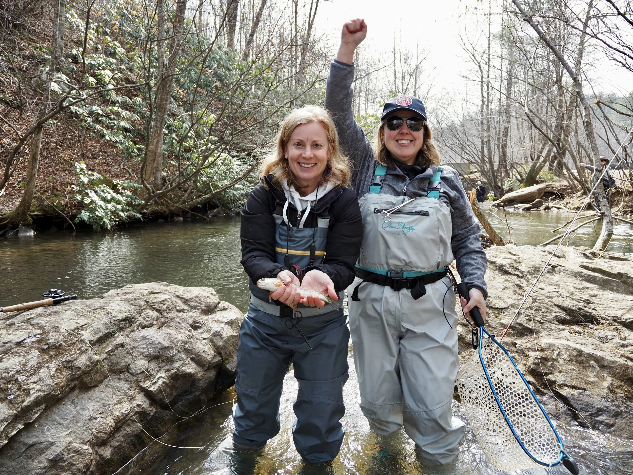 Two women are kneeling by a creek in a wooded area, smiling and holding a small fish, with fishing gear nearby.