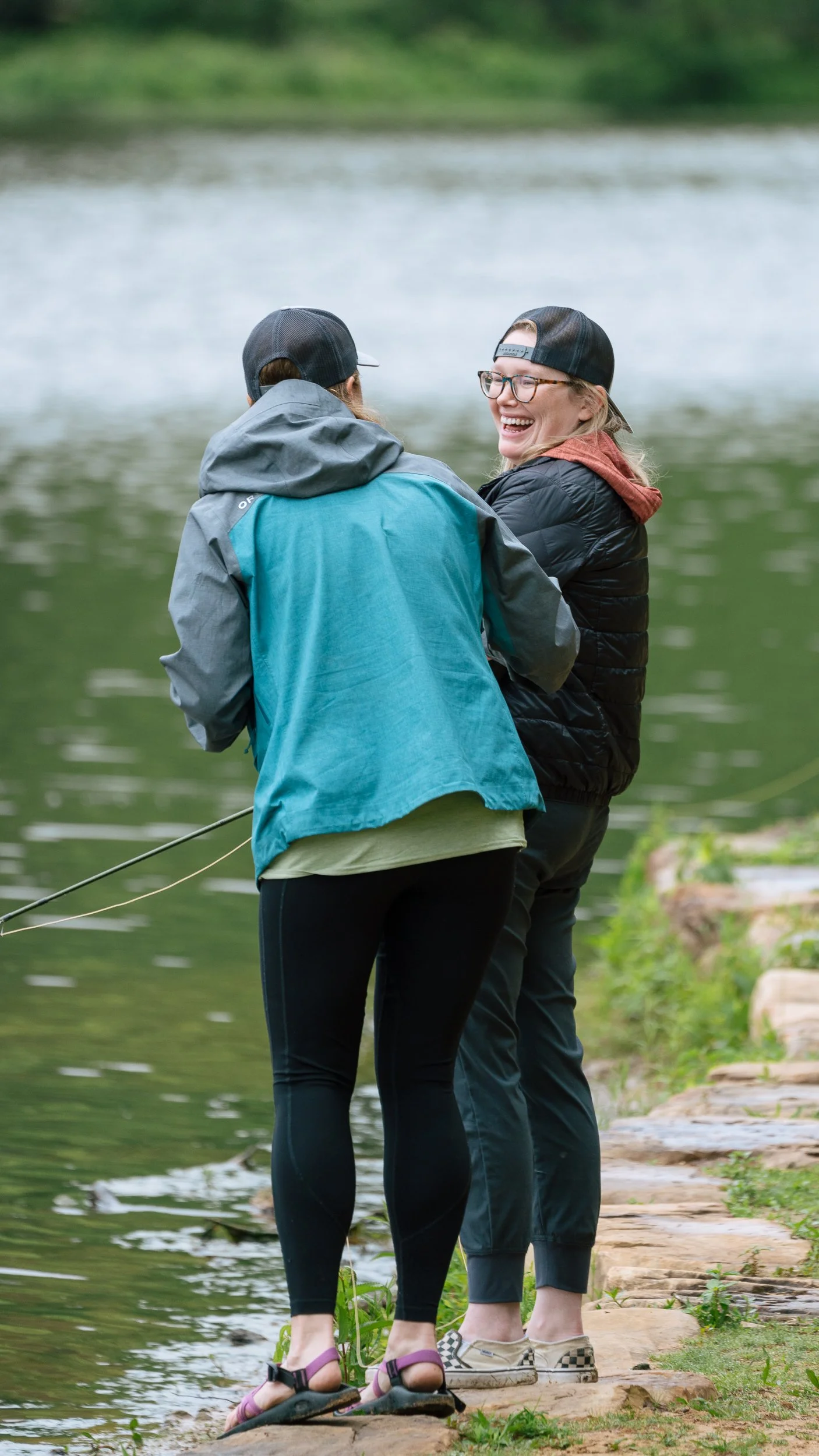 Two women standing on a stone path by a lake, fishing and smiling at each other.