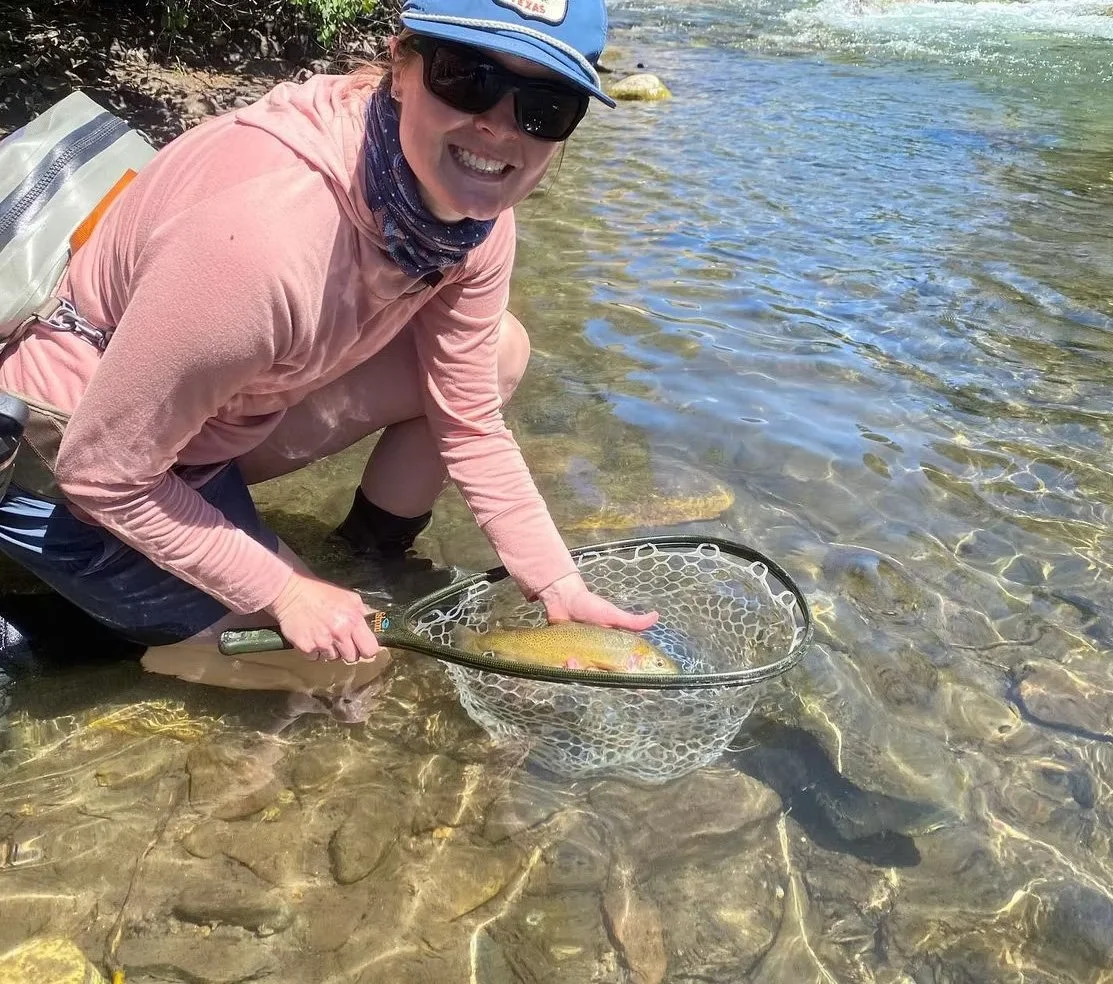 Person in sunglasses, a baseball cap, and outdoor clothing kneels in shallow water holding a fishing net with a large fish inside.