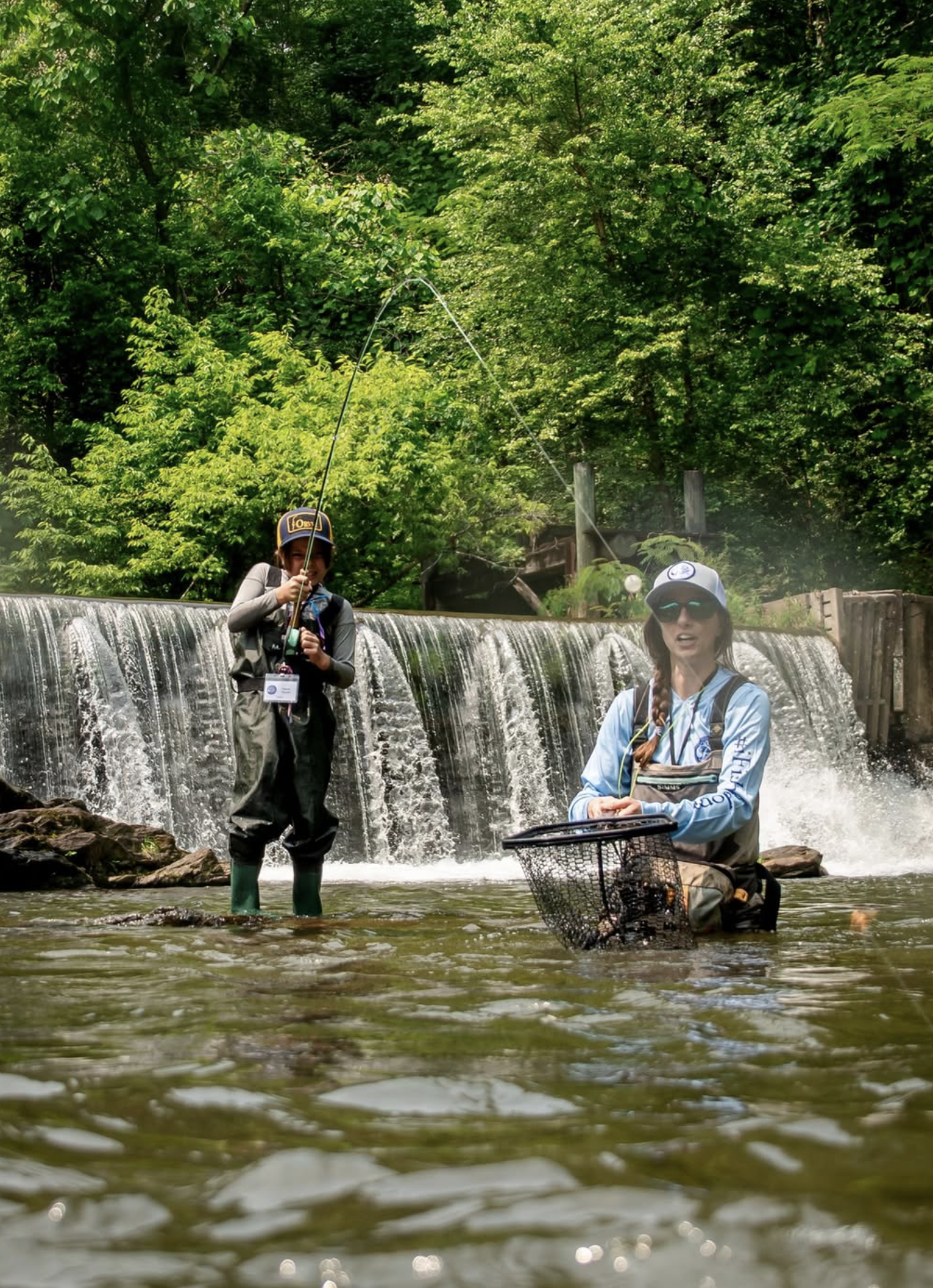 Two women fishing in a river near a small waterfall, surrounded by green trees.