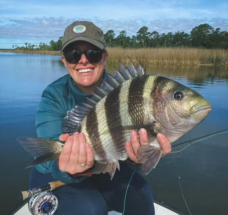 Woman holding a large fish on a boat in a calm water body, with reeds and trees in the background, wearing sunglasses and a hat.